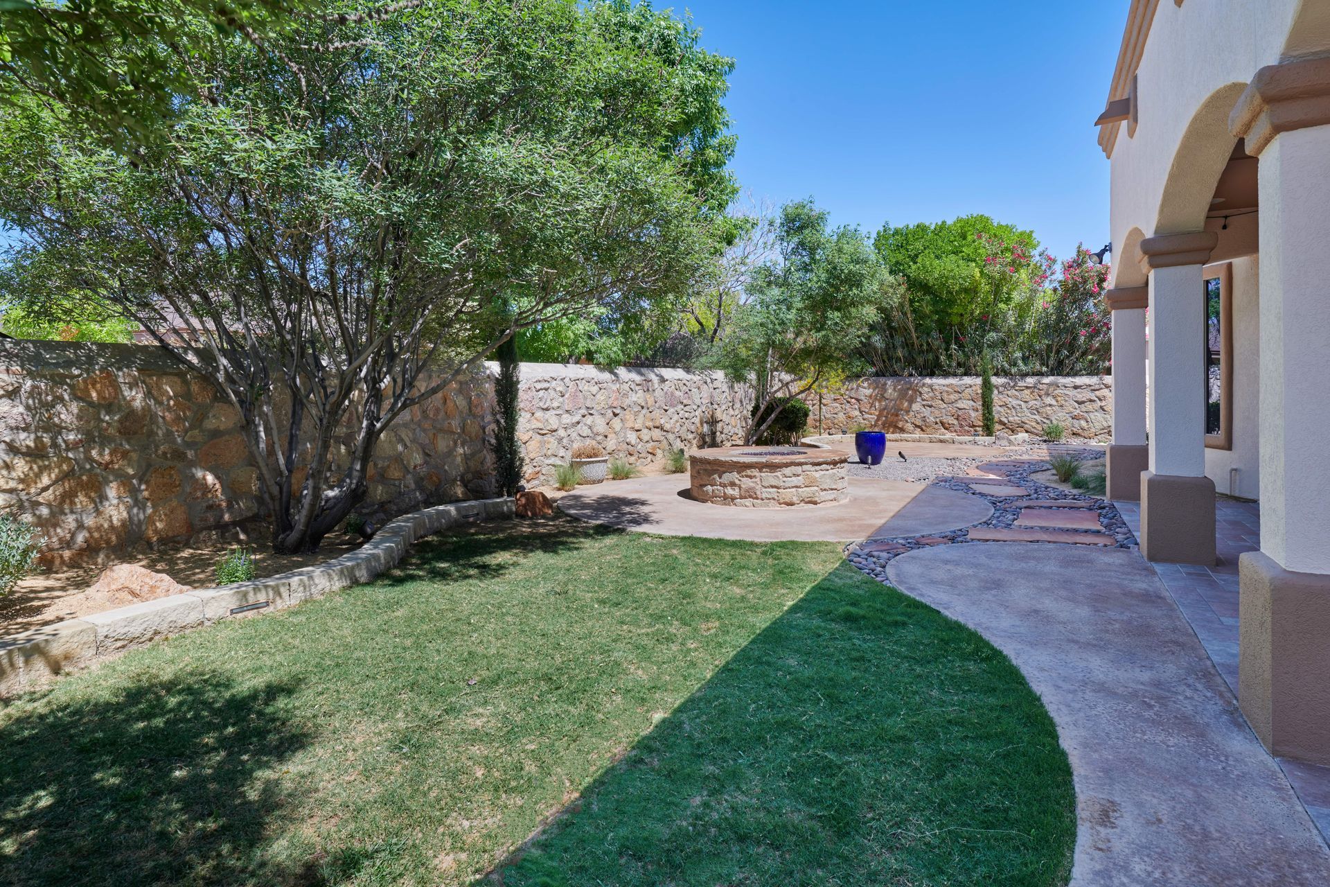 Lawn, stone wall, and patio in a sunny backyard with trees, a blue planter, and a portion of a building.