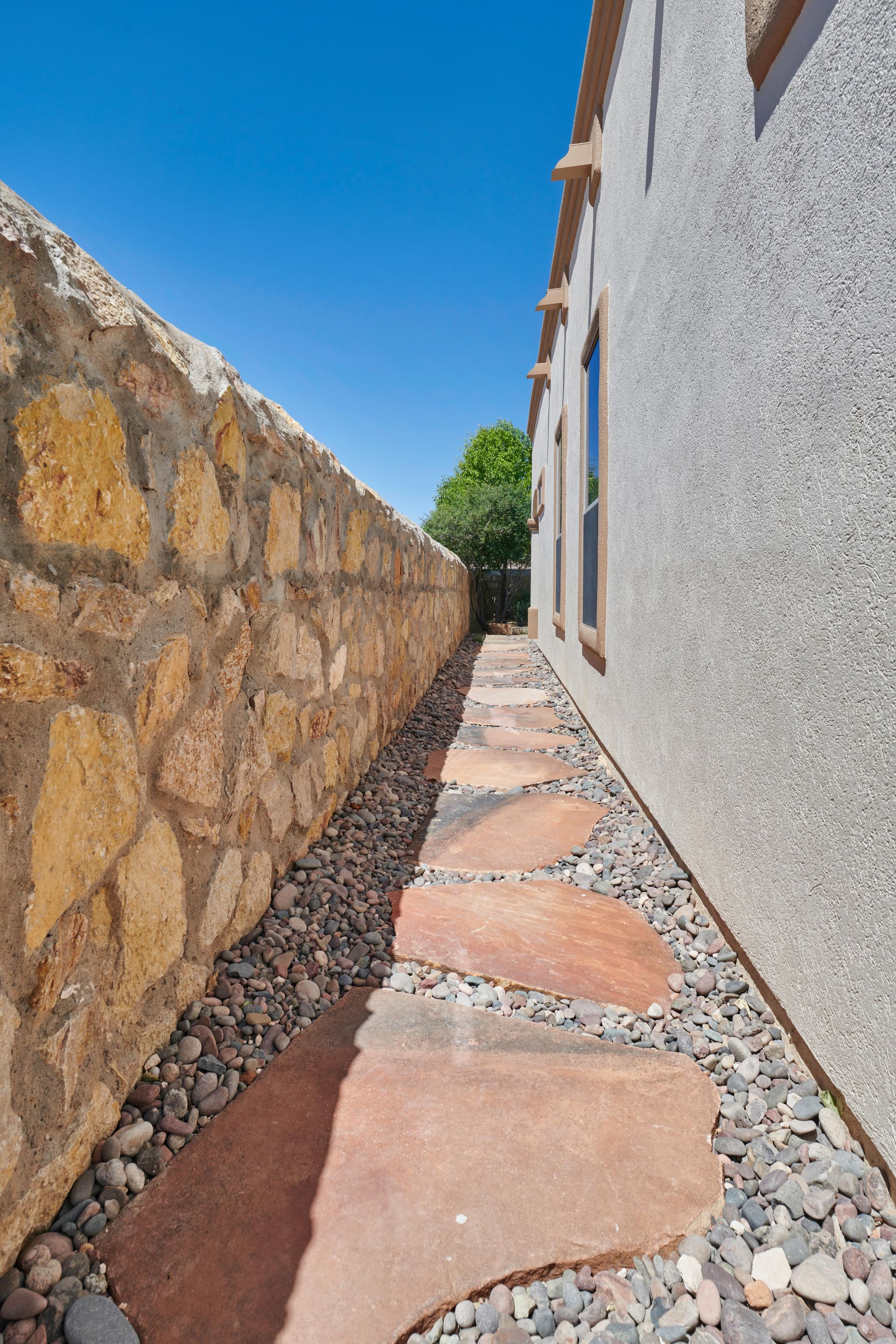 Narrow walkway with stone pavers, bordered by a stone wall and building with windows, under a blue sky.