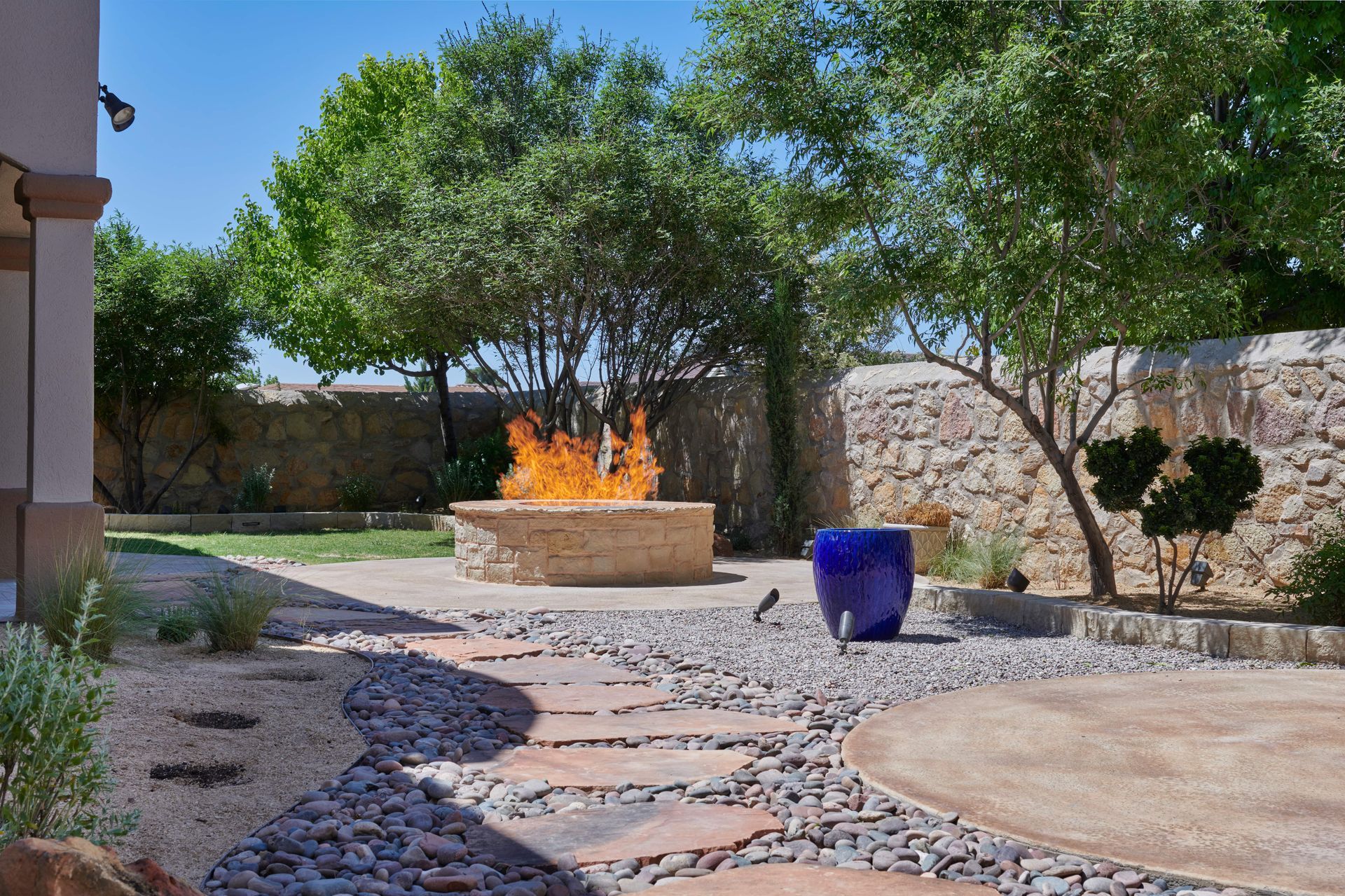 Backyard with stone pathway, fire pit with flames, blue pot, and green trees.