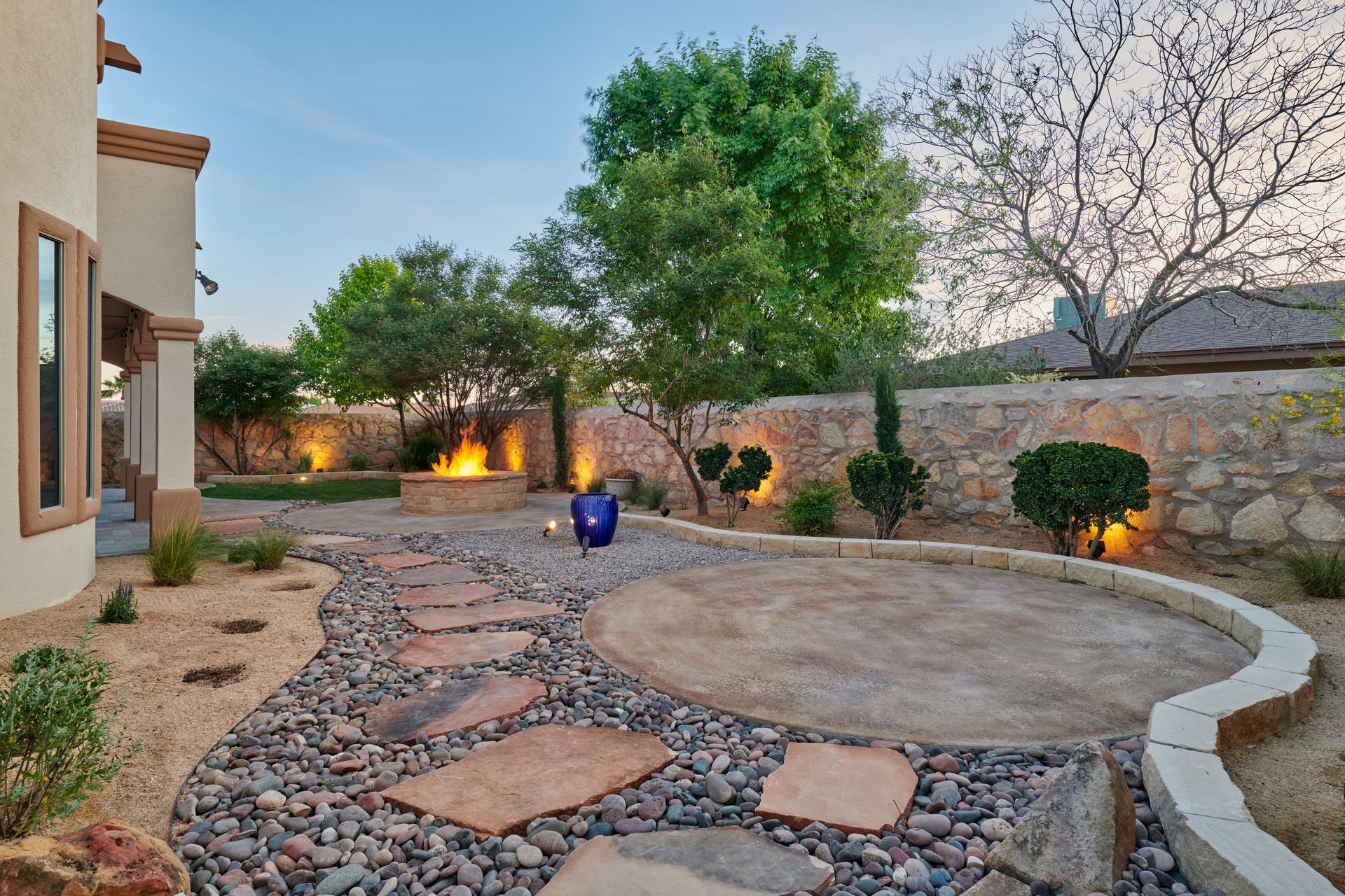 Backyard landscape with fire pit, stone path, patio, and desert plants at dusk.