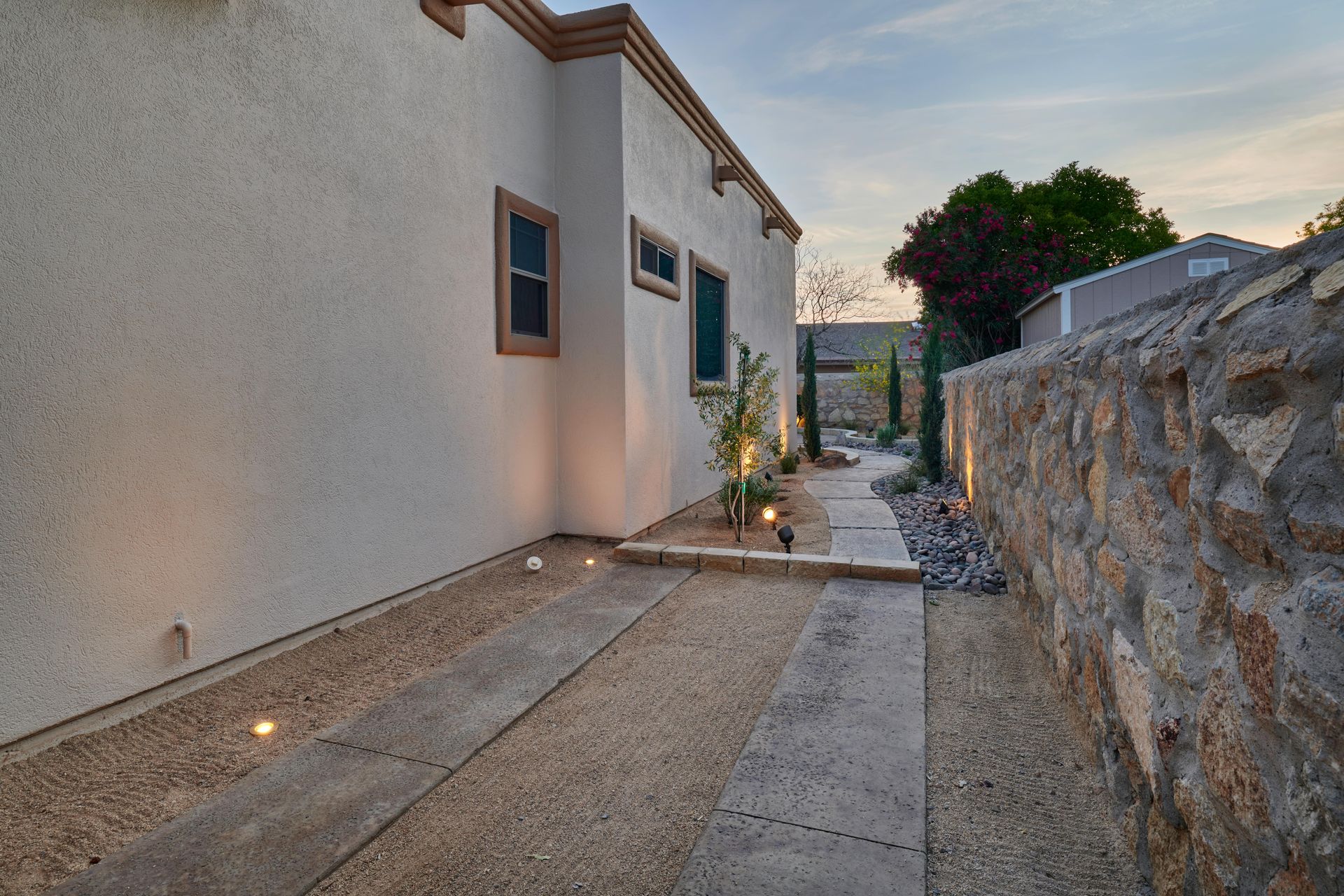 Exterior shot of a building with a textured wall, pathway, and rock wall; pathway is illuminated.