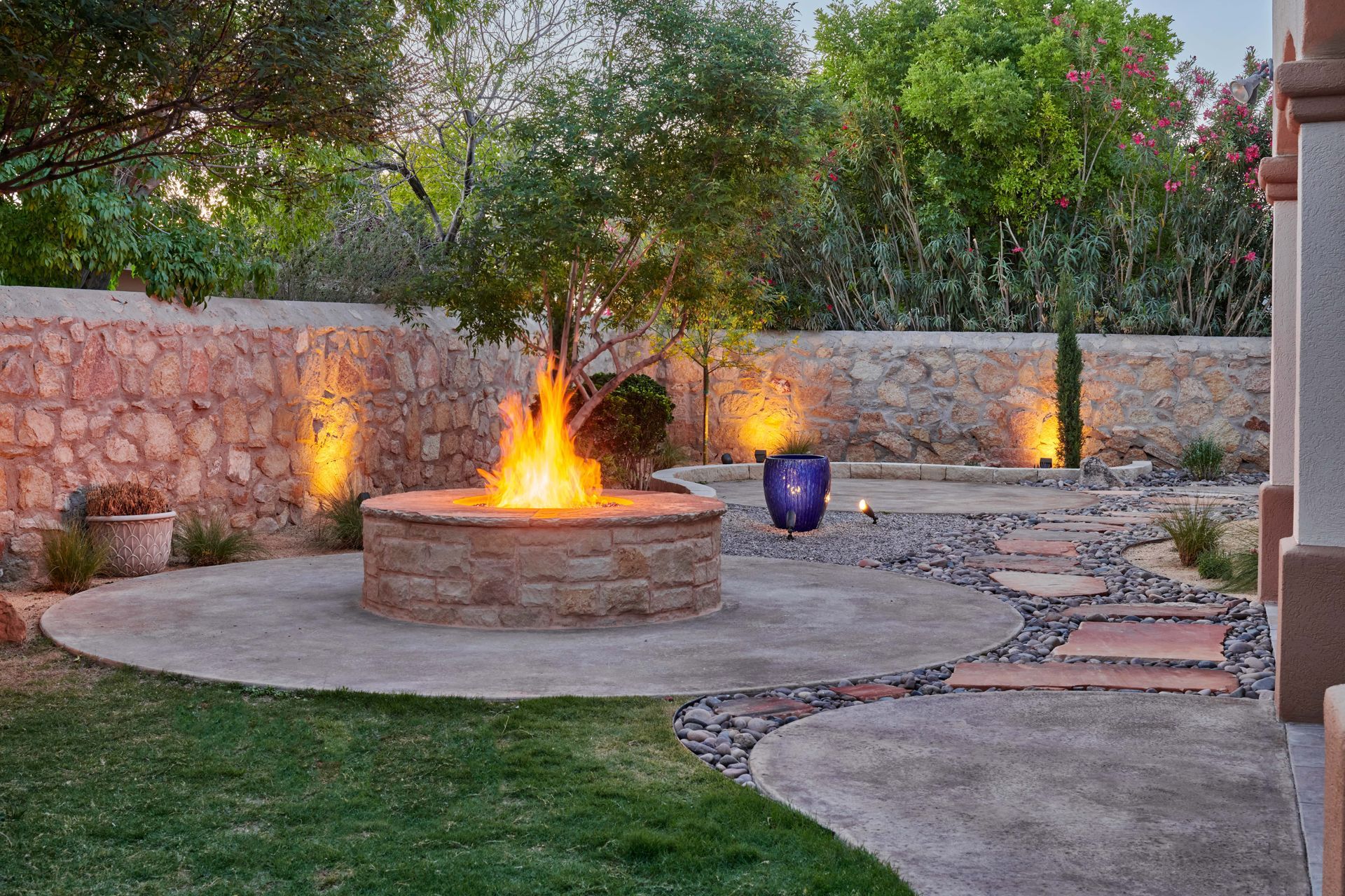Fire pit with flames, surrounded by stone patio, landscaping, and outdoor lighting.