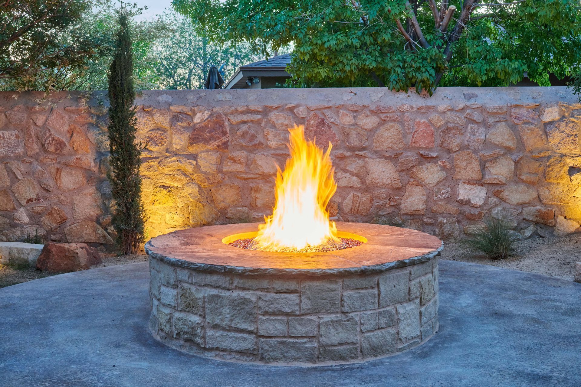 Fire pit with flames, surrounded by a stone wall. Flames glowing brightly. Evening setting.