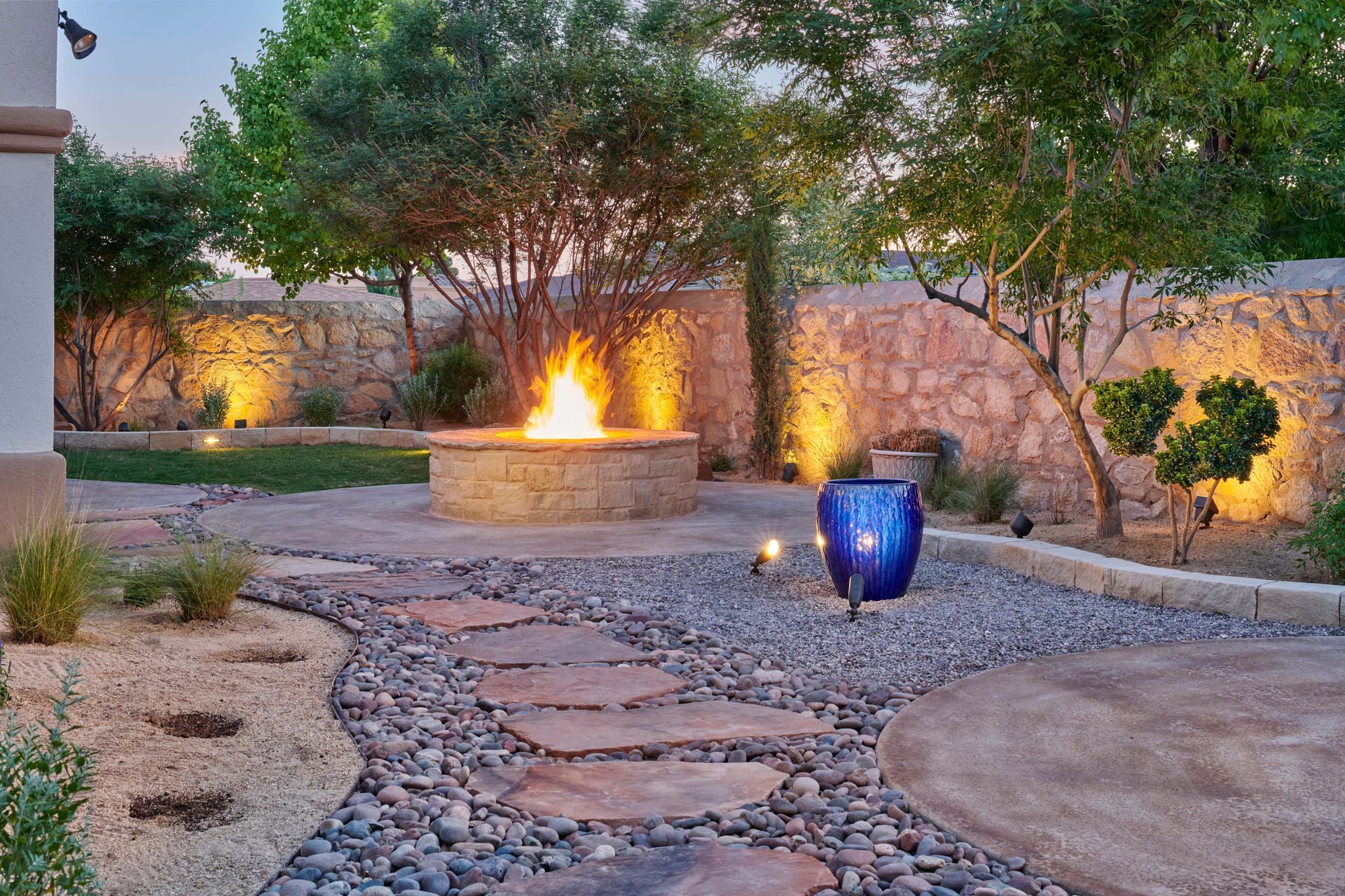 Stone patio with fire pit, illuminated trees, and pathway, creating a warm, inviting backyard setting.