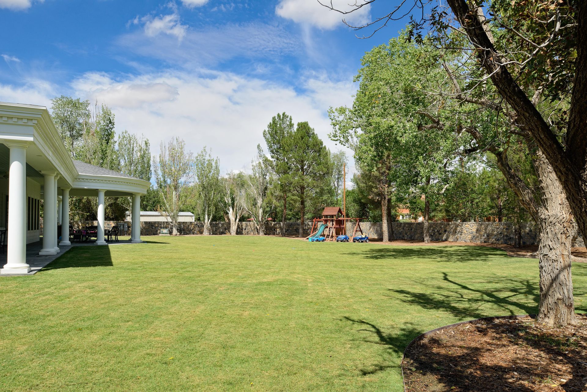 Lawn in backyard with covered porch on the left, trees in the background, and a playset in the distance.