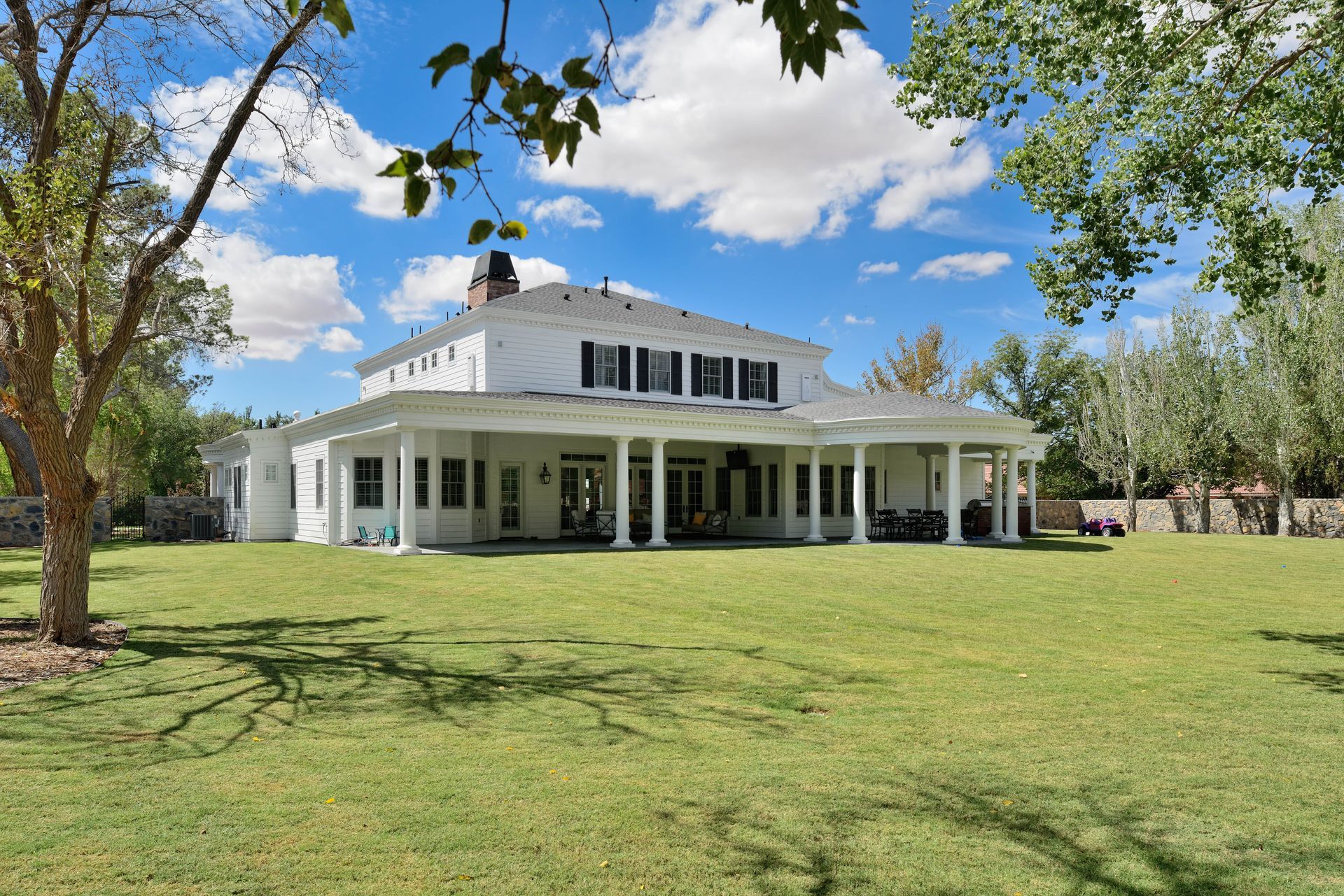 White two-story house with black shutters, large porch, and green lawn under a bright, cloudy sky.