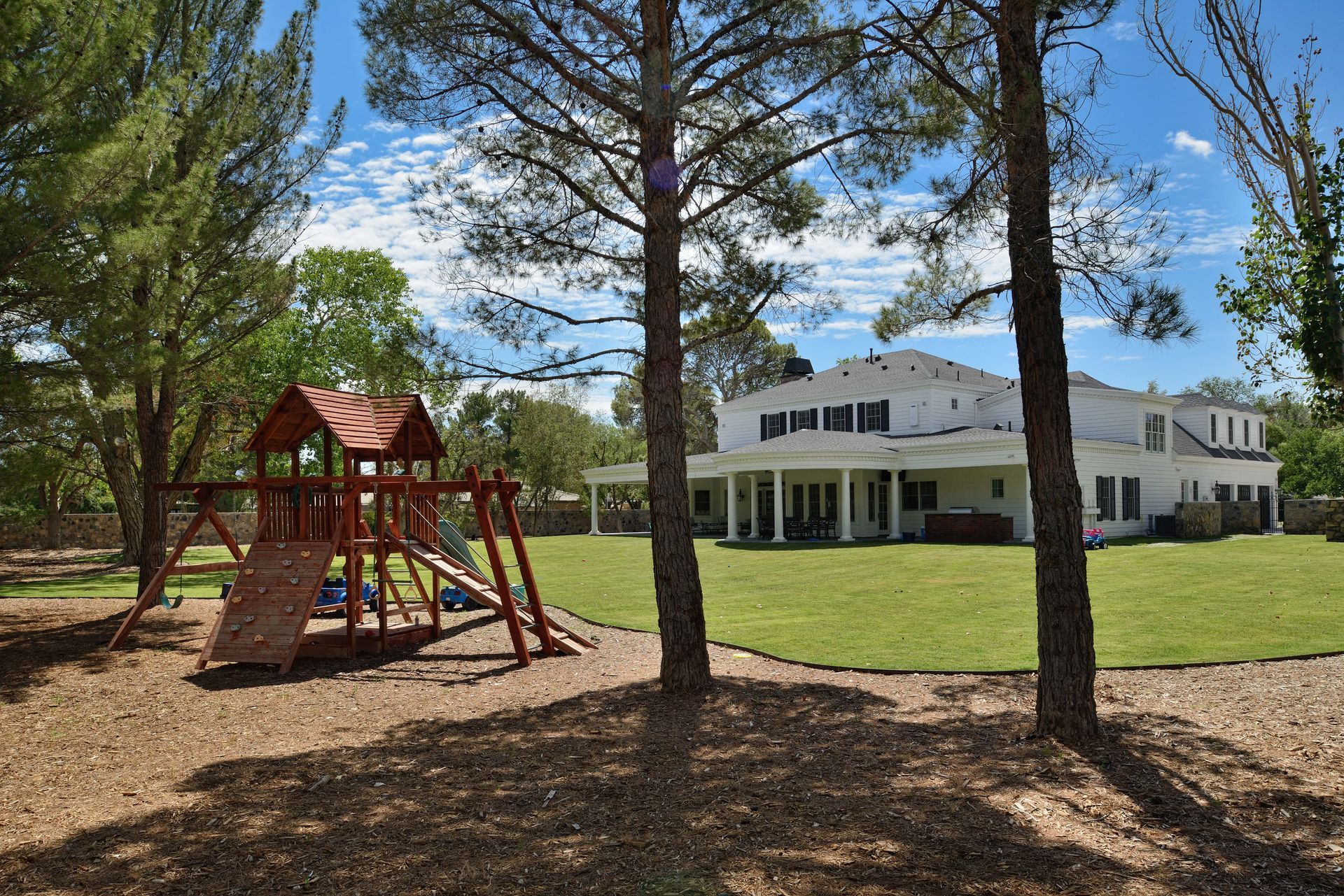 Playground in front of a white house with a green lawn. Trees frame the view under a blue sky.