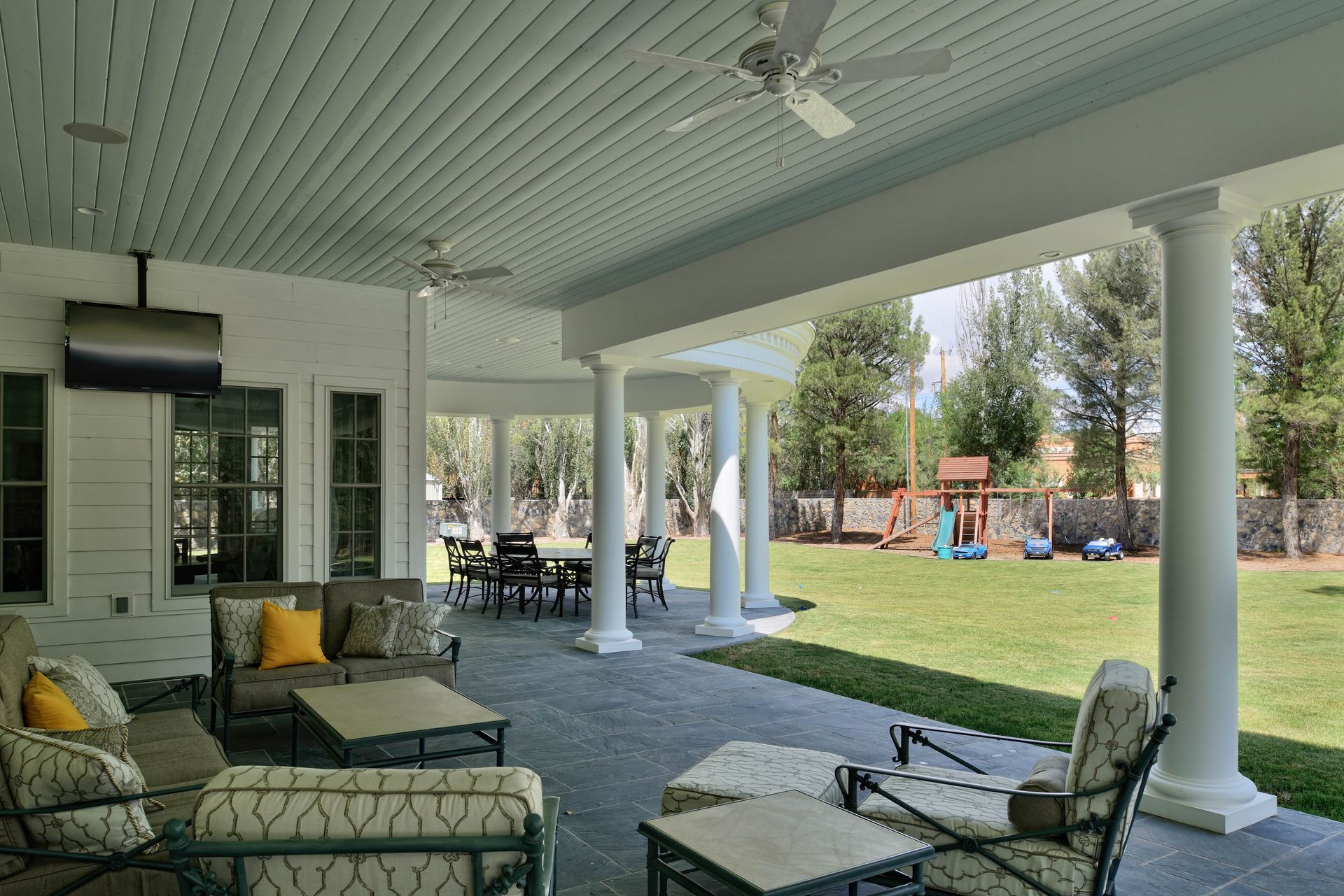 Covered patio with seating, television, and columns overlooking a backyard with a playground.