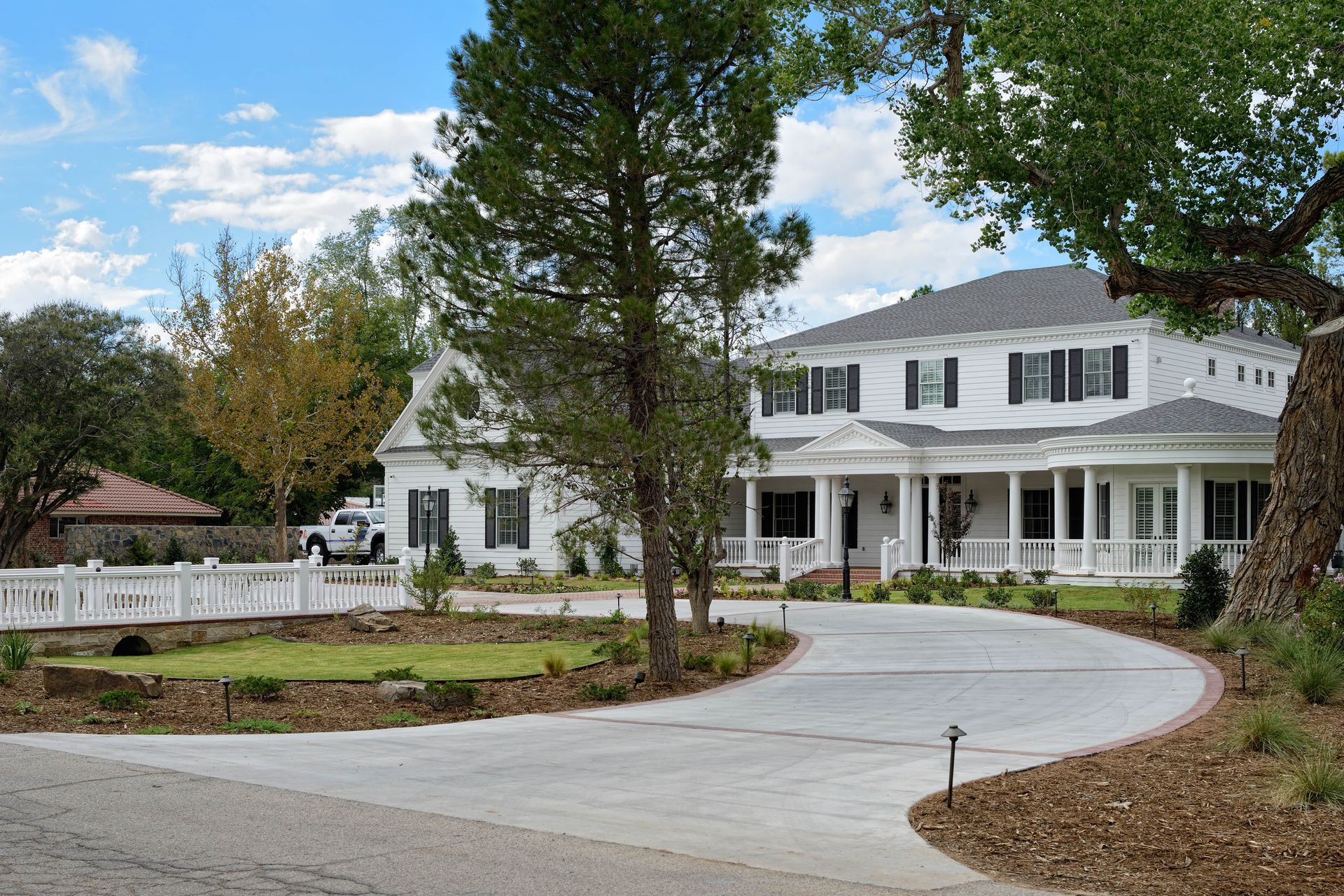 White two-story house with black shutters, circular driveway, and small detached building on a sunny day.