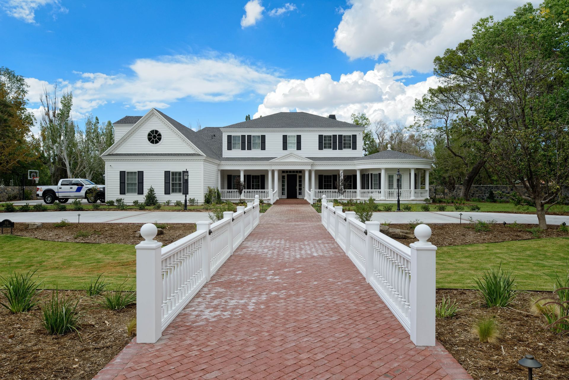 White two-story house with black shutters, long red brick walkway, and green lawn under a blue sky.