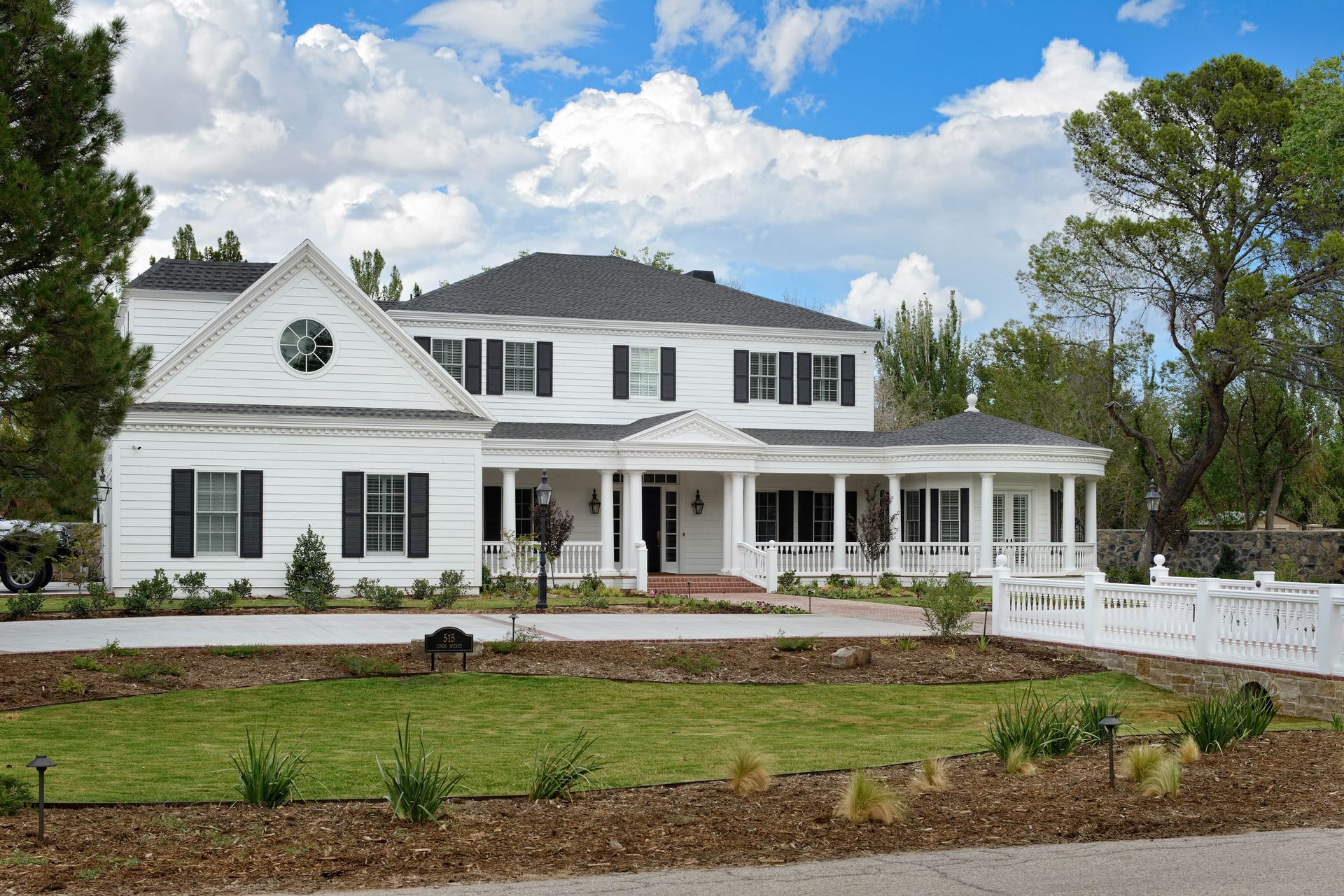 White two-story house with black shutters and a covered porch. Sunny day, green lawn, brown mulch beds.