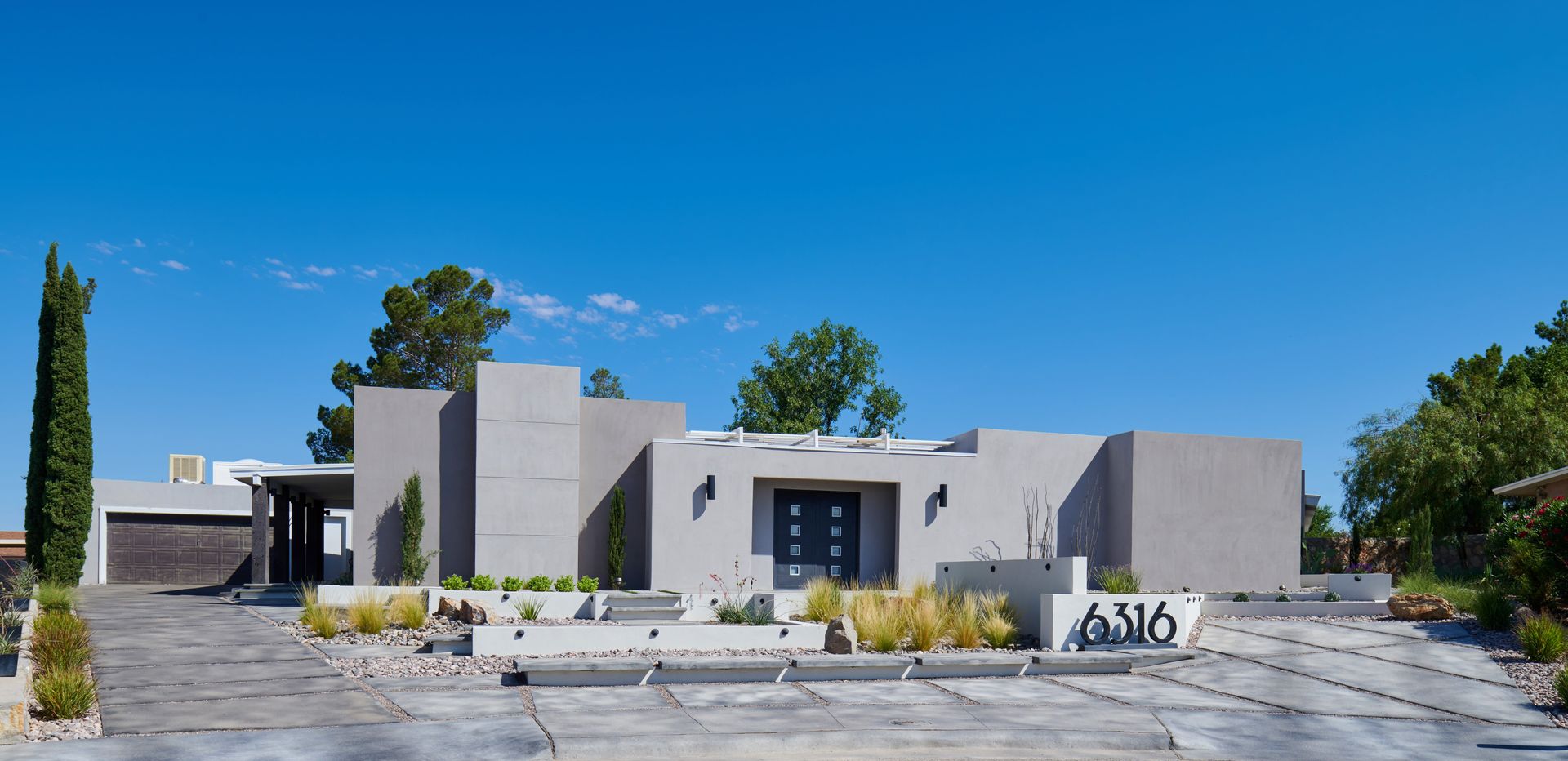 Modern, gray house with a flat roof and a blue sky. Concrete driveway and landscaping in front.