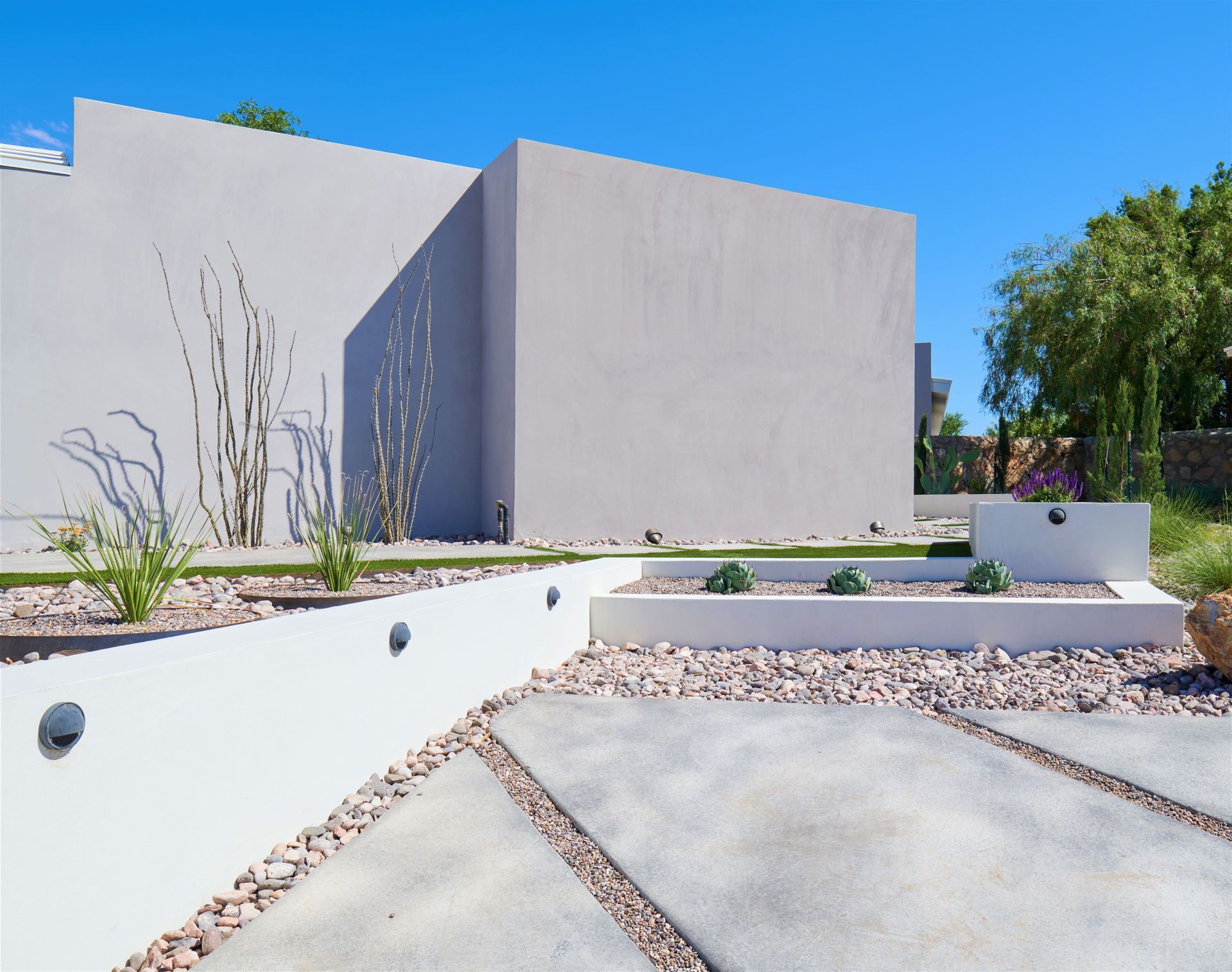 Modern home exterior with gray stucco walls, concrete patio, and desert landscaping under a blue sky.