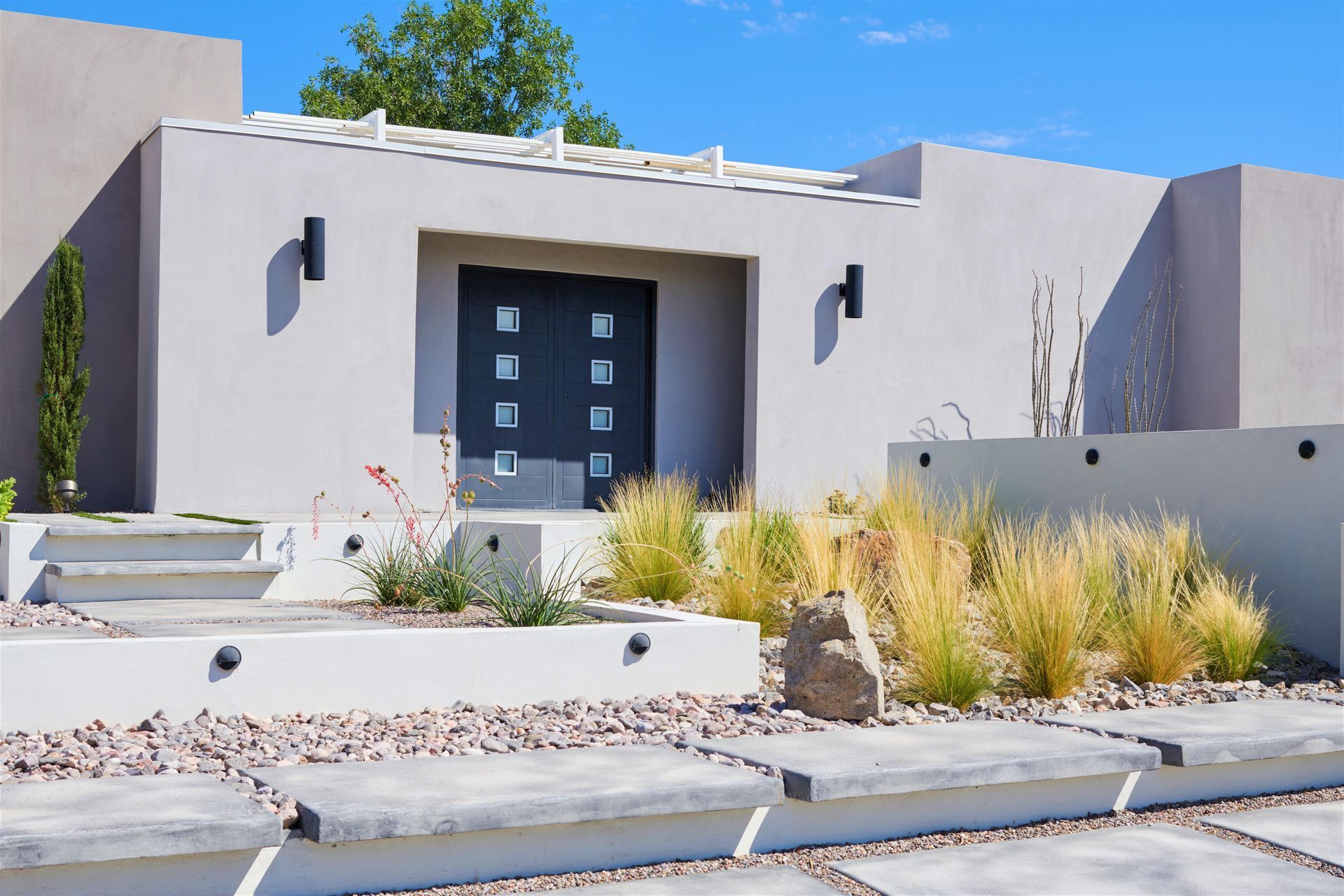 Modern home exterior with gray stucco, steps, landscaping, and a dark front door.