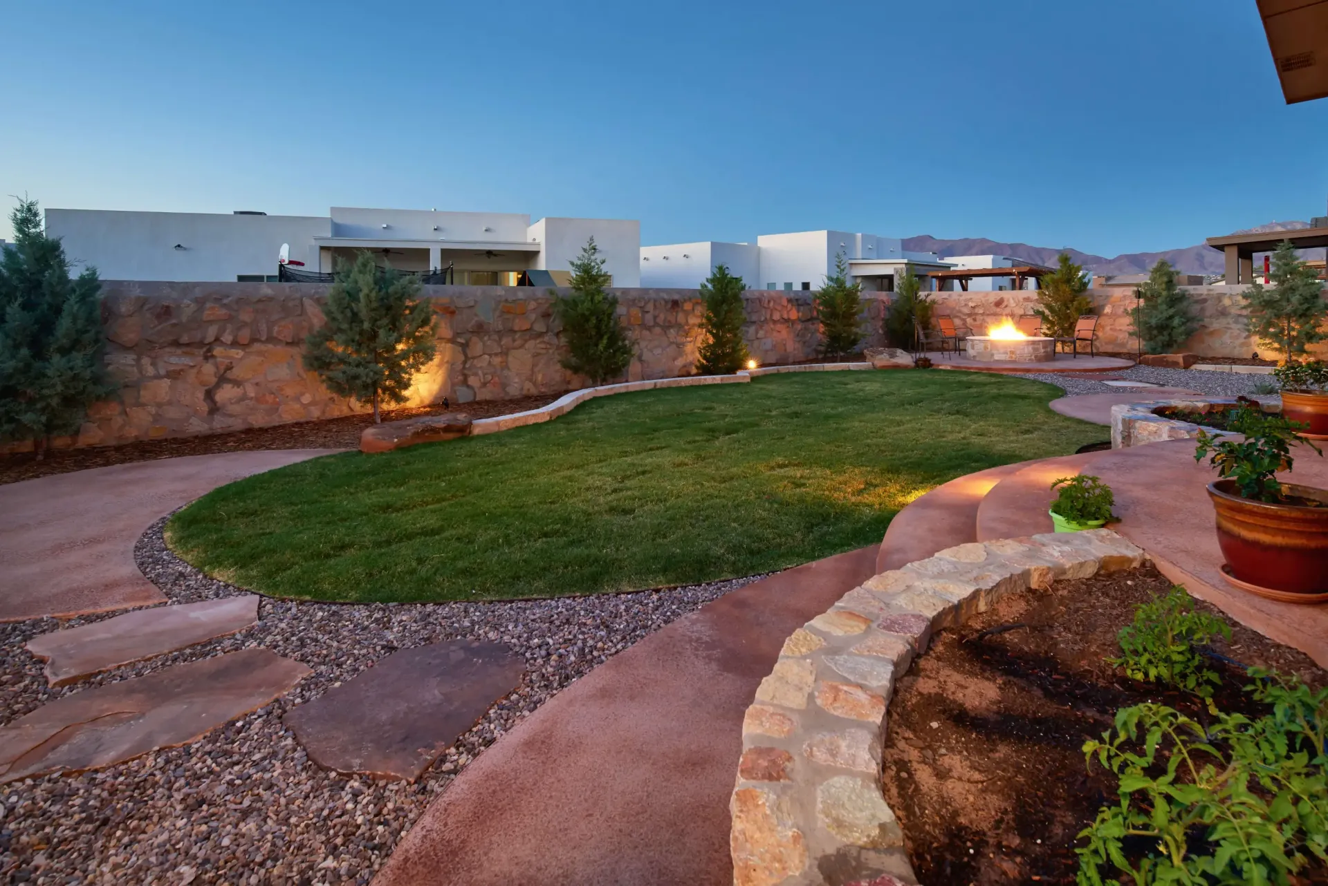 Backyard with green lawn, stone path, landscaping, and a fire pit at dusk.