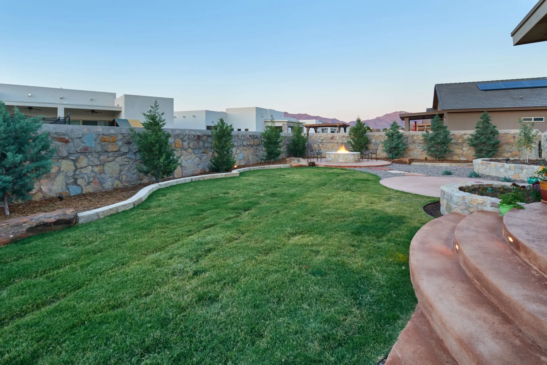 Green lawn in a landscaped backyard with stone walls, trees, and houses in the distance.