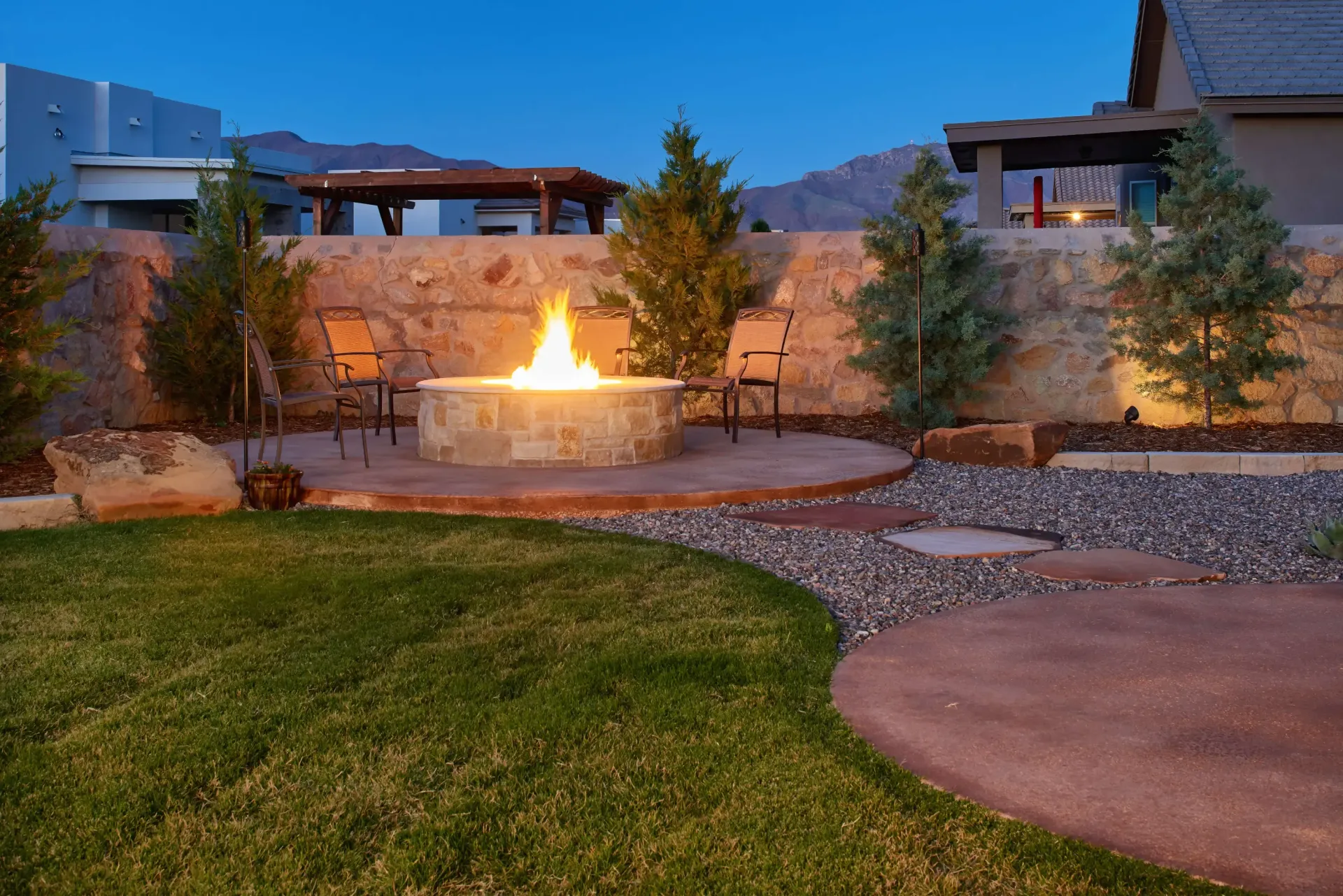 Fire pit with chairs on a patio with grass, gravel, trees, and a stone wall in the evening.