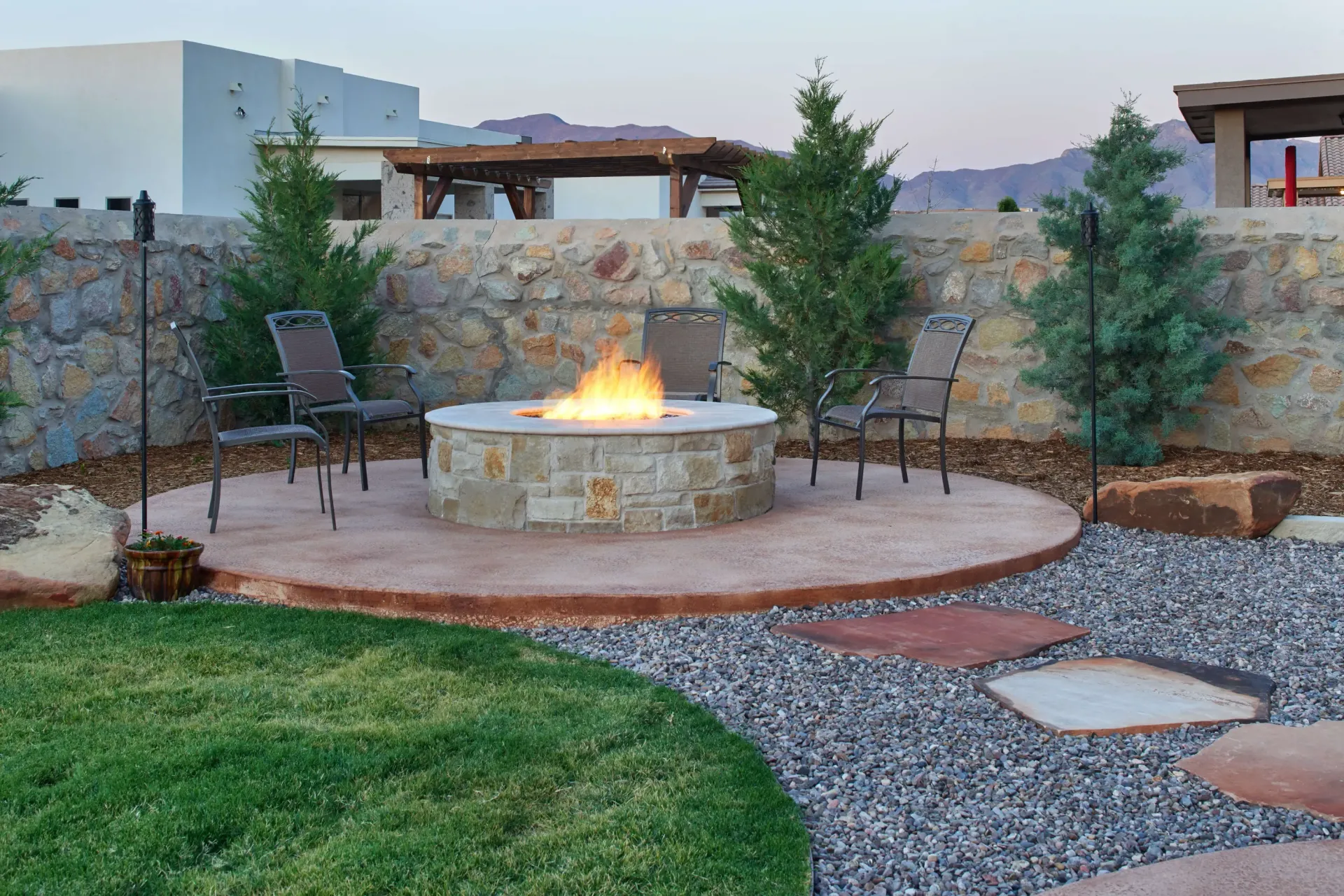 Stone fire pit with two chairs in a backyard setting, surrounded by a stone wall, plants, and gravel pathway.