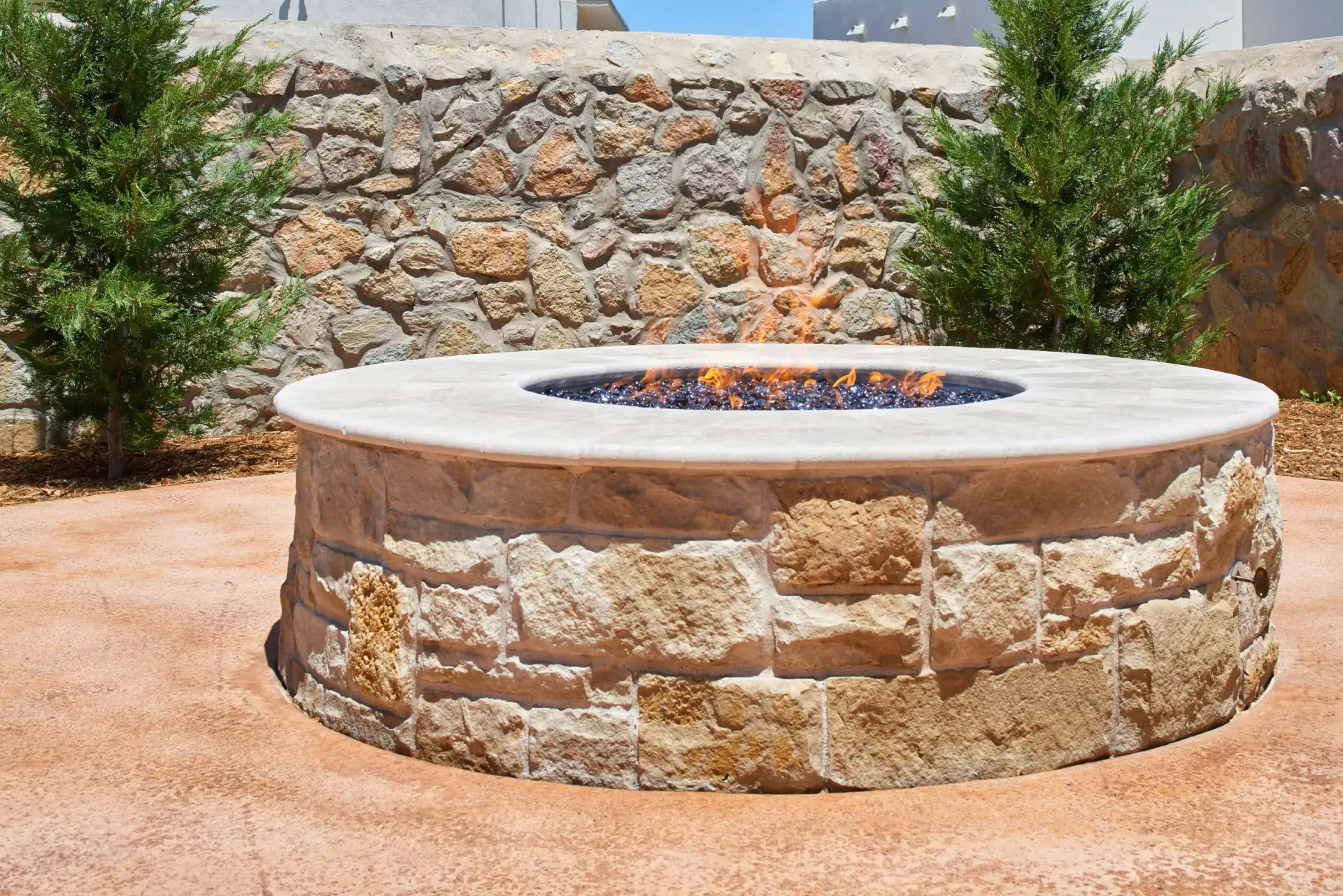 Round stone fire pit with flames, in a tan gravel setting, with a stone wall and green bushes.