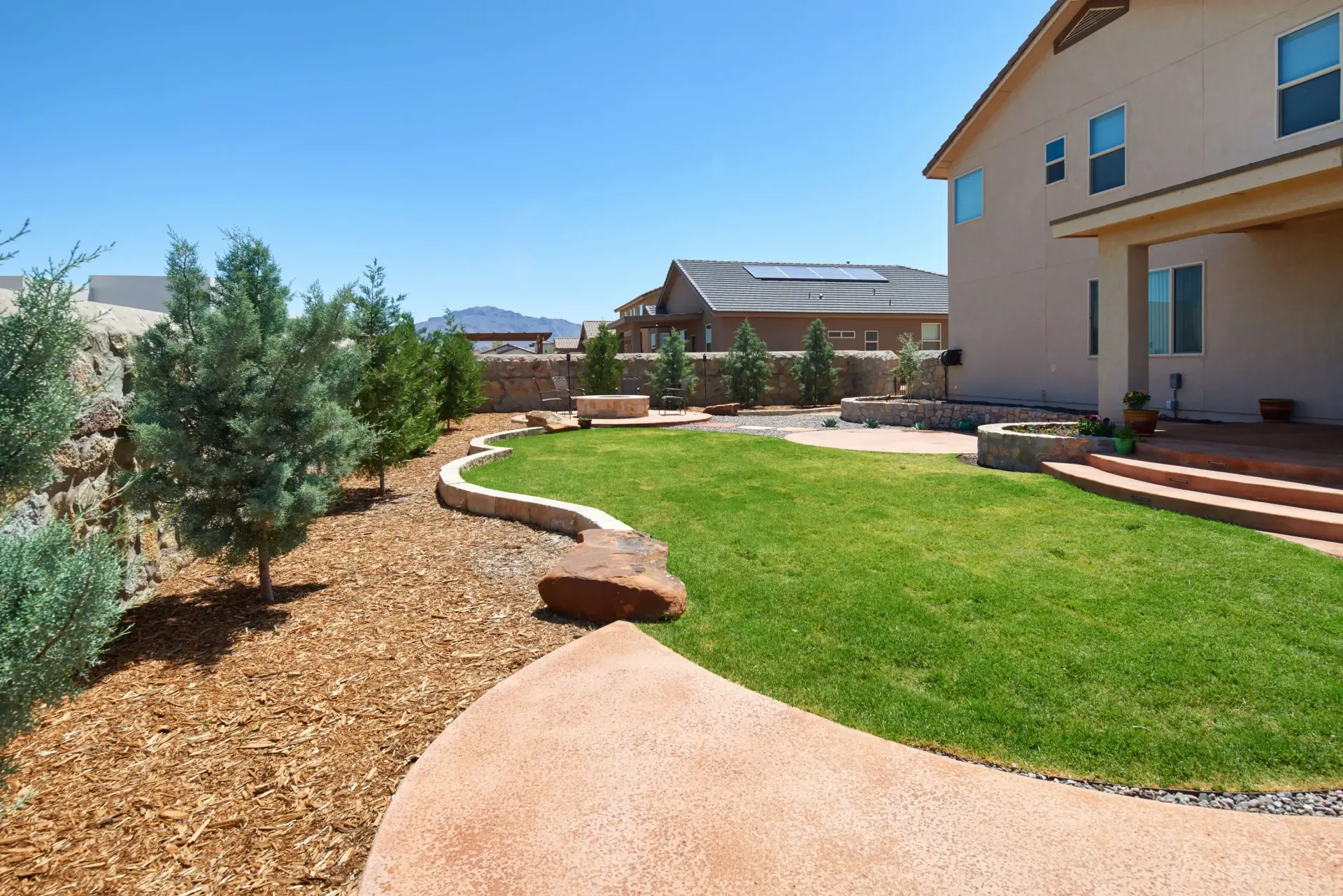 Lush green backyard with stone pathway, raised garden bed, house, and blue sky.