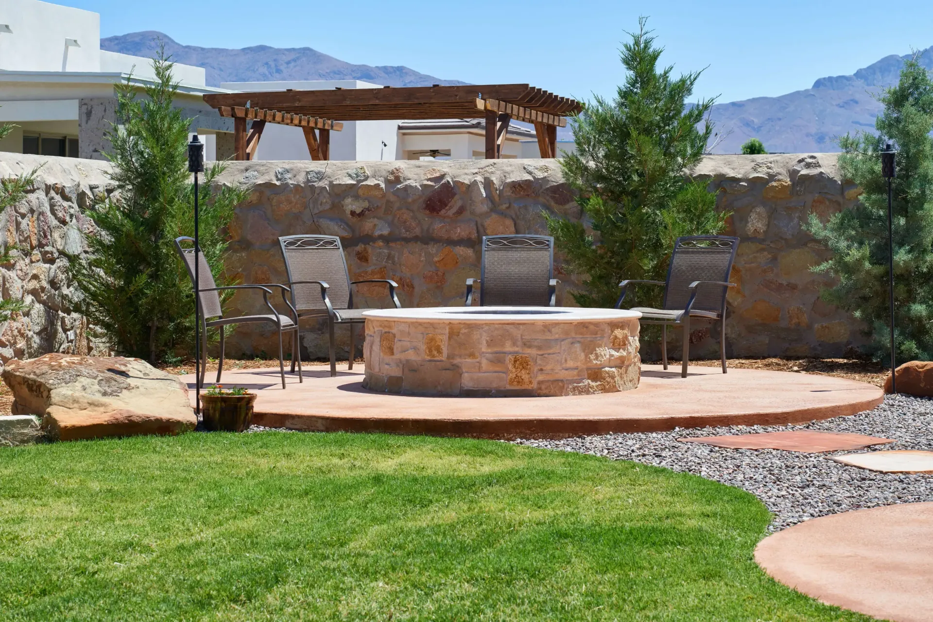 Fire pit with chairs on a circular stone patio, surrounded by green grass and a stone wall.