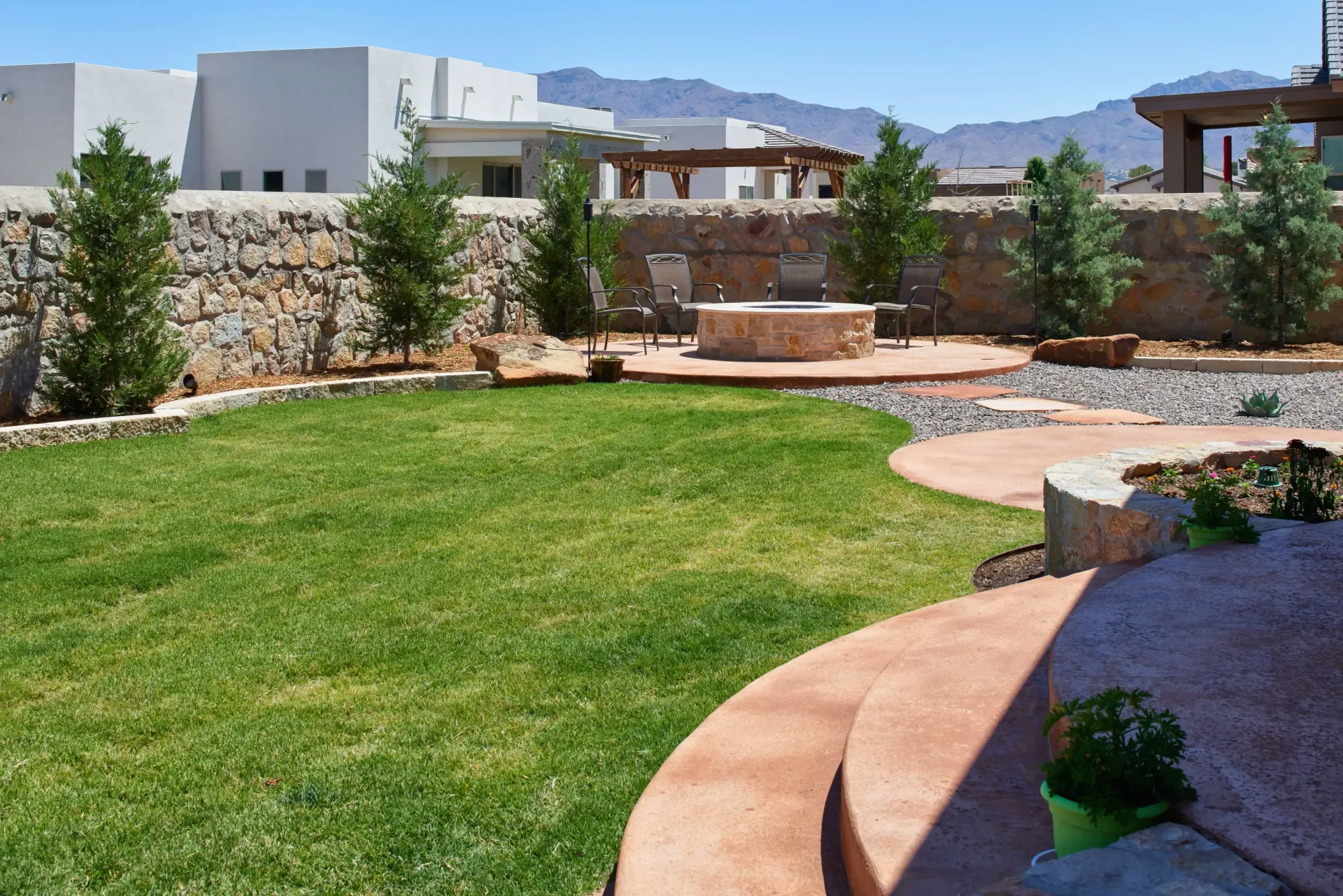 Backyard with green lawn, stone fire pit, and rock wall under a blue sky.