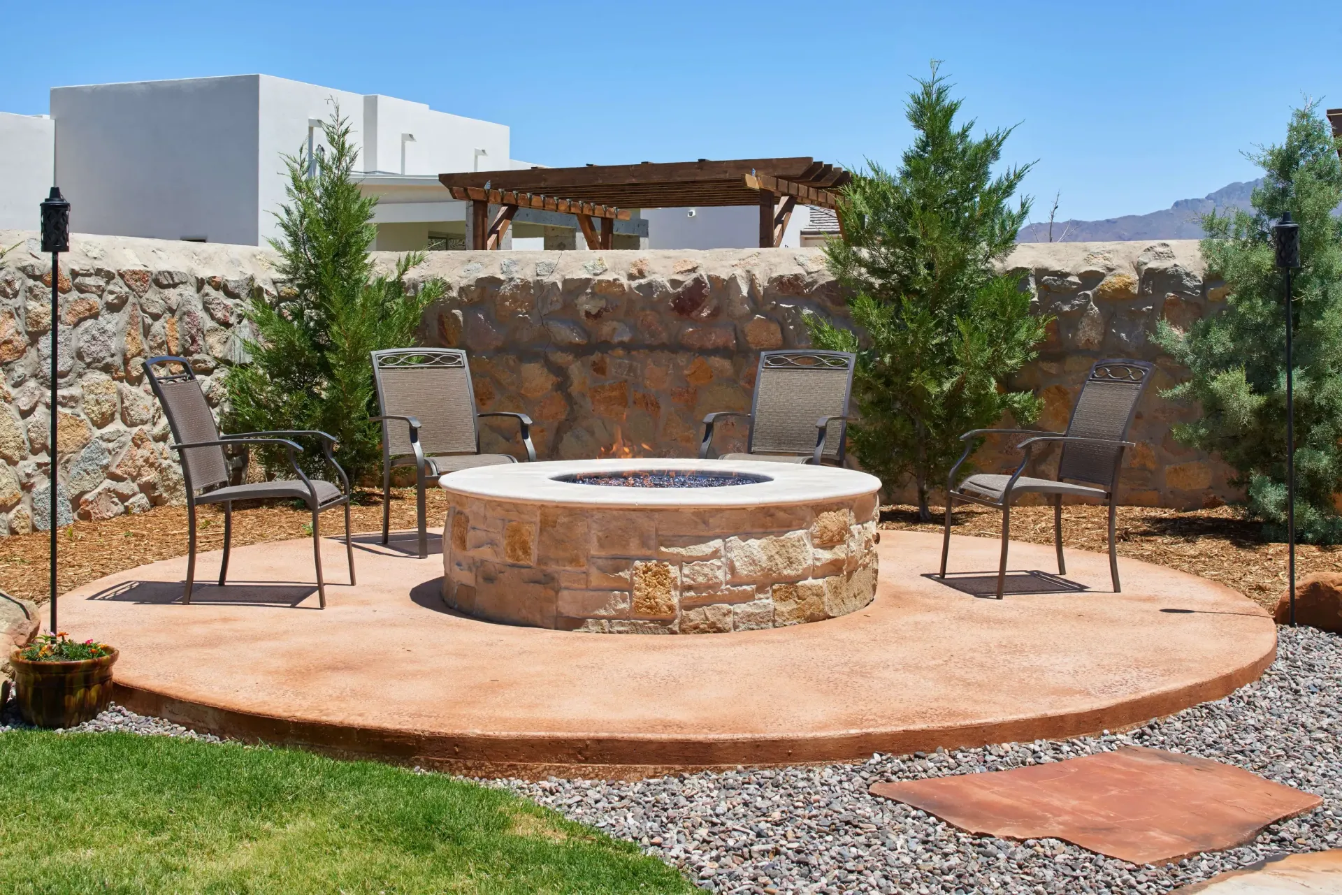 Outdoor fire pit with chairs, surrounded by gravel and trees, under a blue sky.