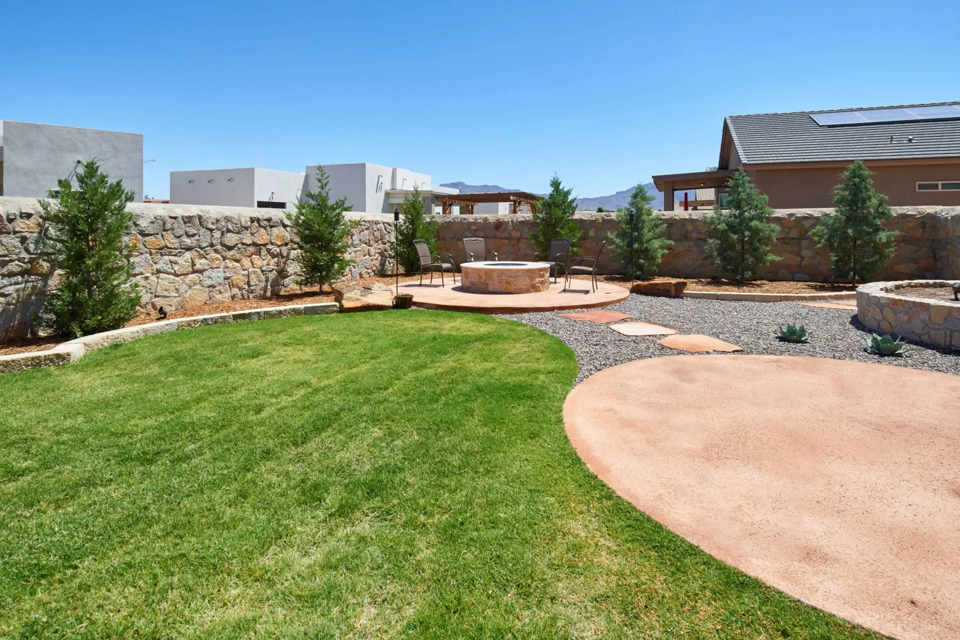 Backyard with green grass, stone fire pit, and a red patio under a clear blue sky.