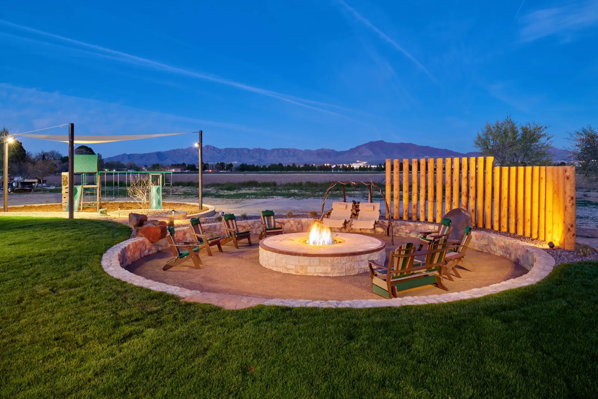 Fire pit area with seating, wooden fence, grass lawn, mountains, and dusk sky.