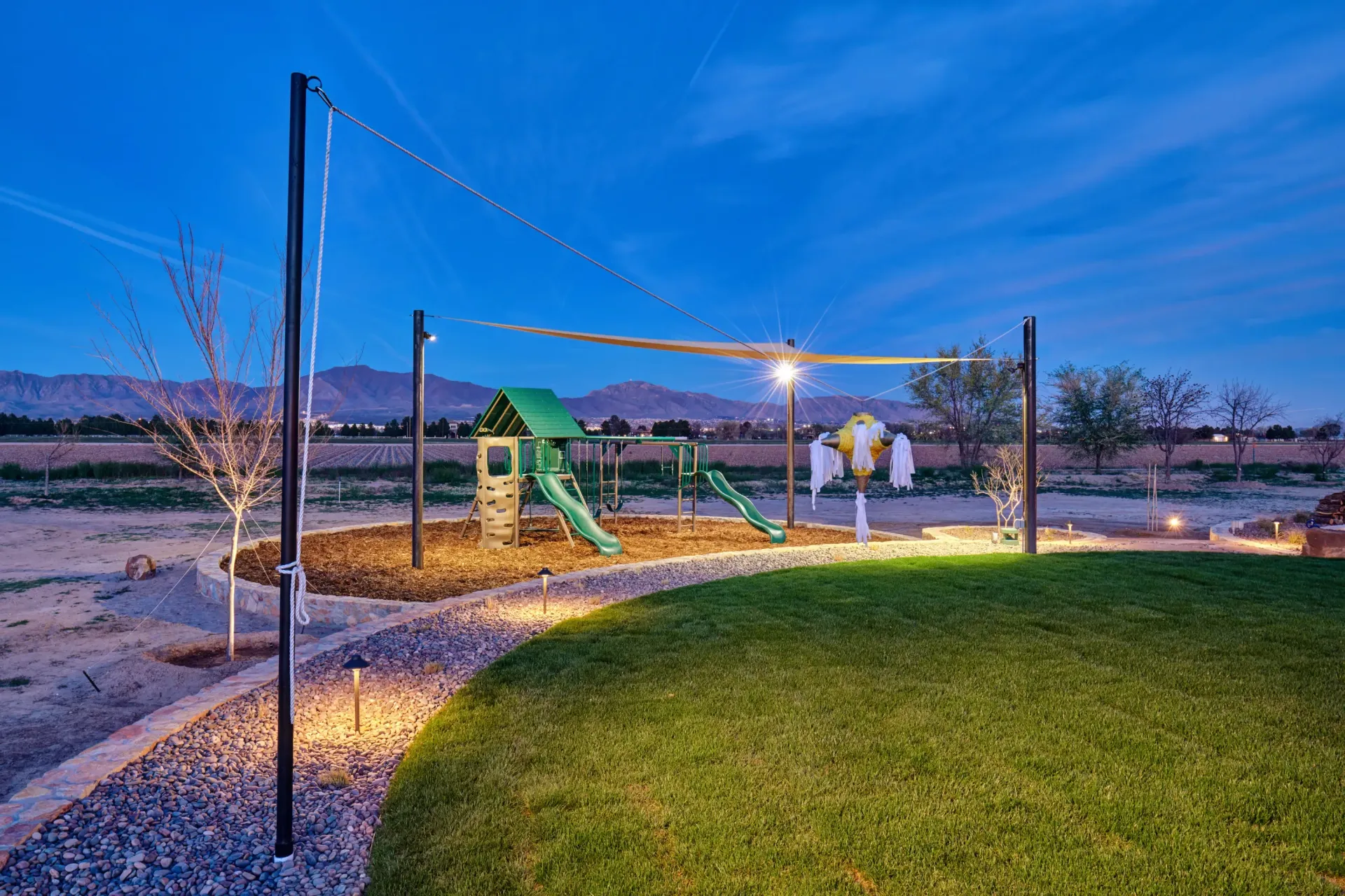 Playground at dusk, lit by string lights and spotlights. Includes a slide, swing set, and shade sail.