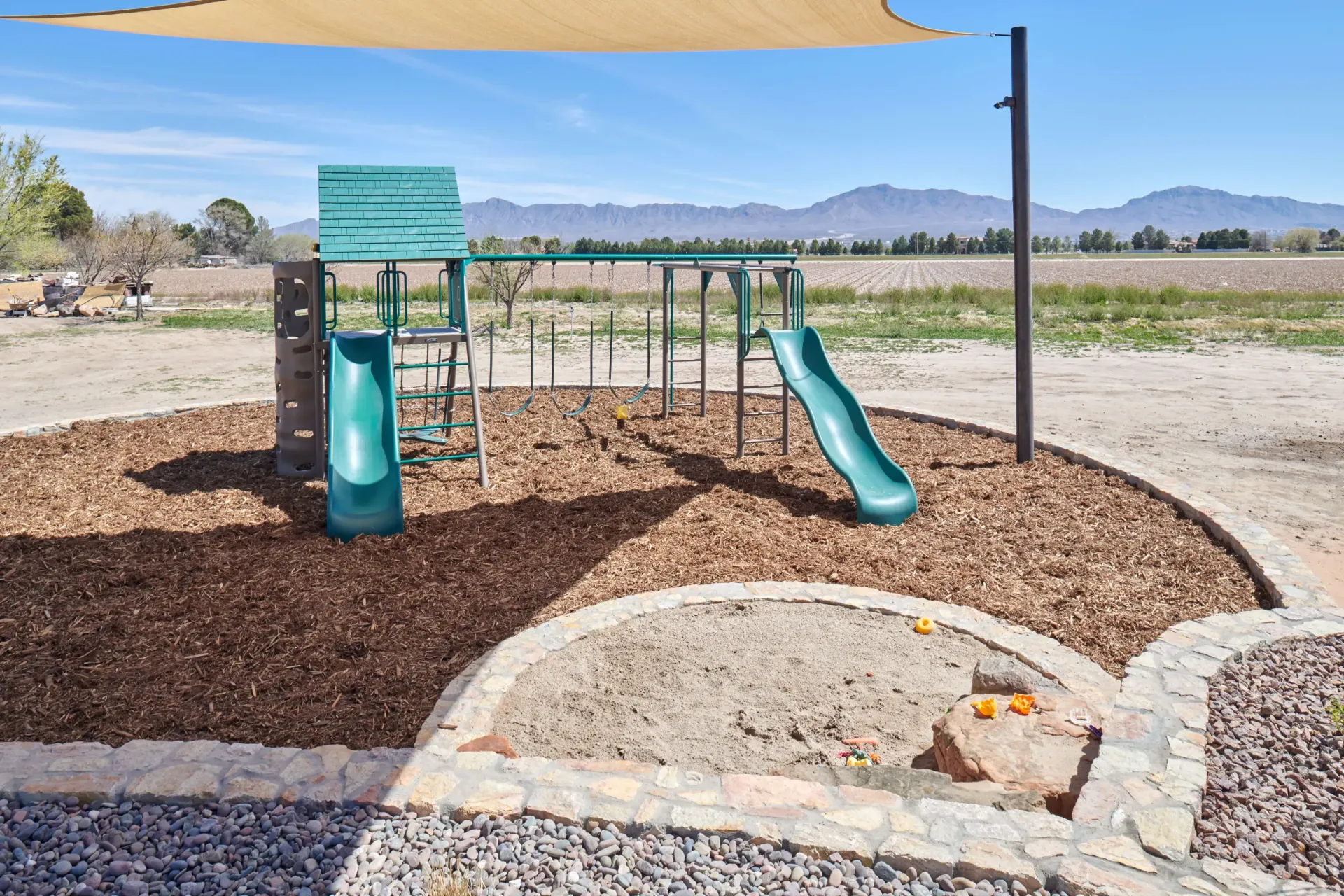 Playground with slides, climbing wall, and monkey bars, set in a wood-chip area under a sunshade, with mountains in the background.