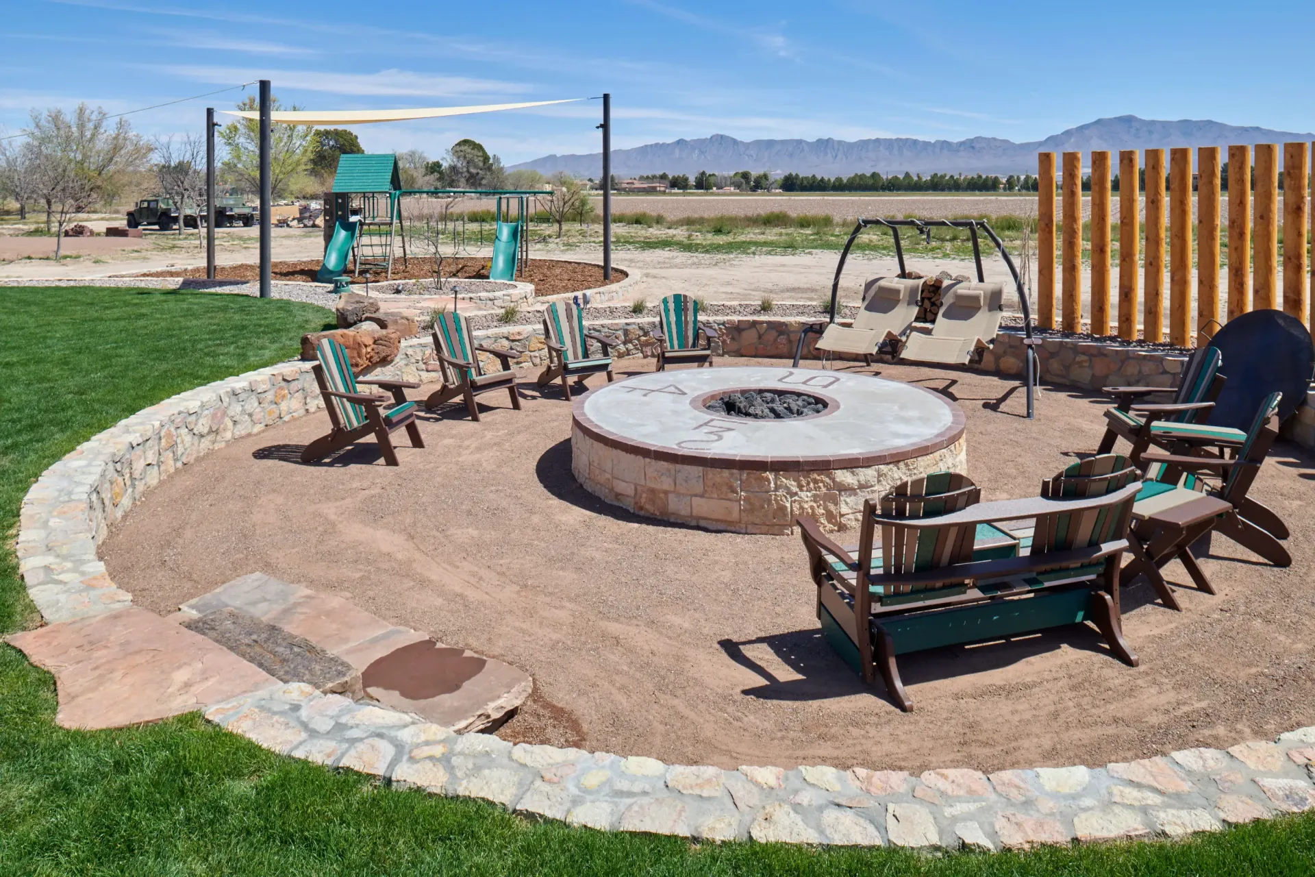 Outdoor fire pit with seating, surrounded by gravel, green grass, and play set, against a rural landscape.