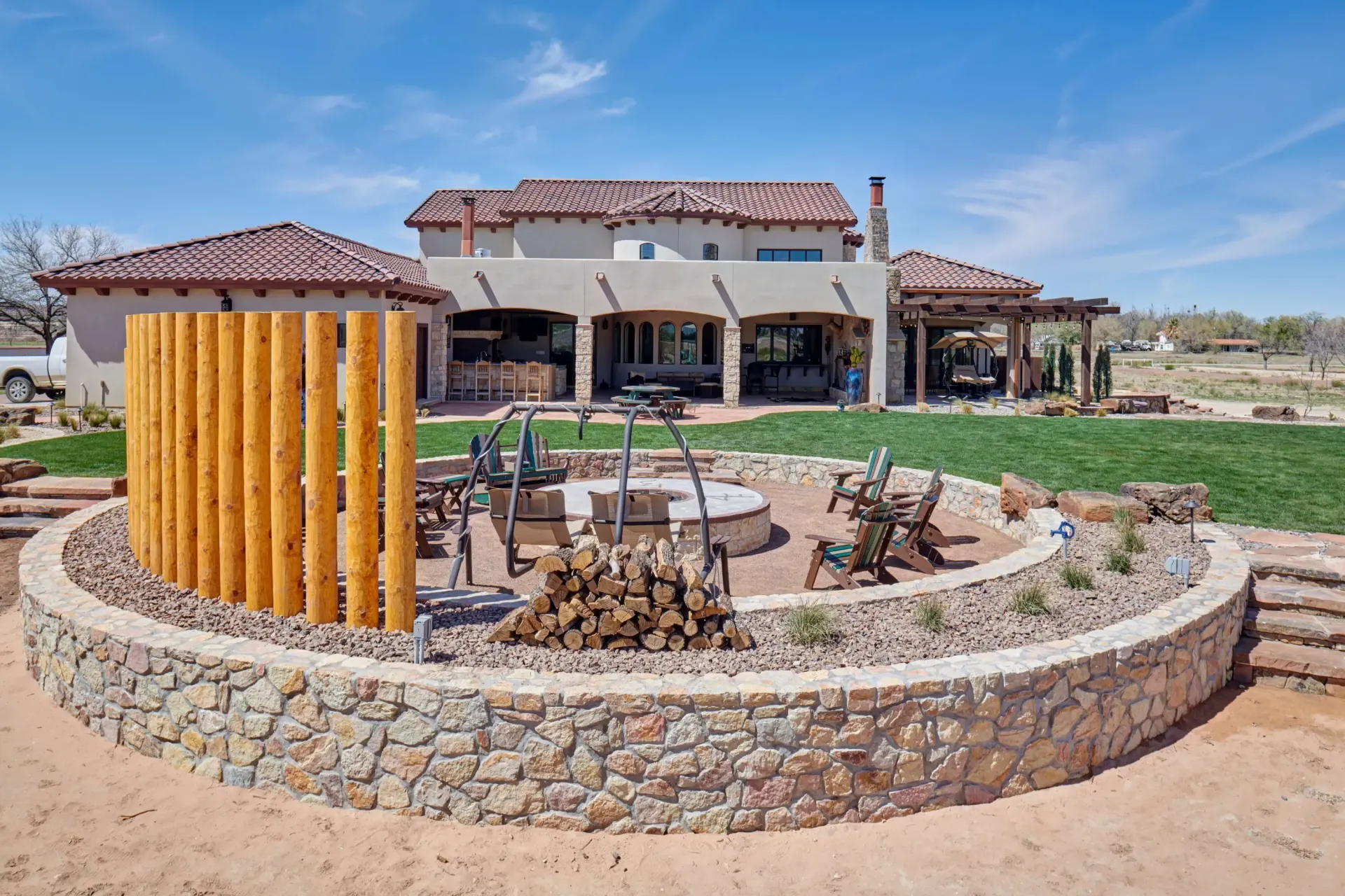 Backyard fire pit with stone wall and log privacy screen, overlooking a large house with a tile roof.