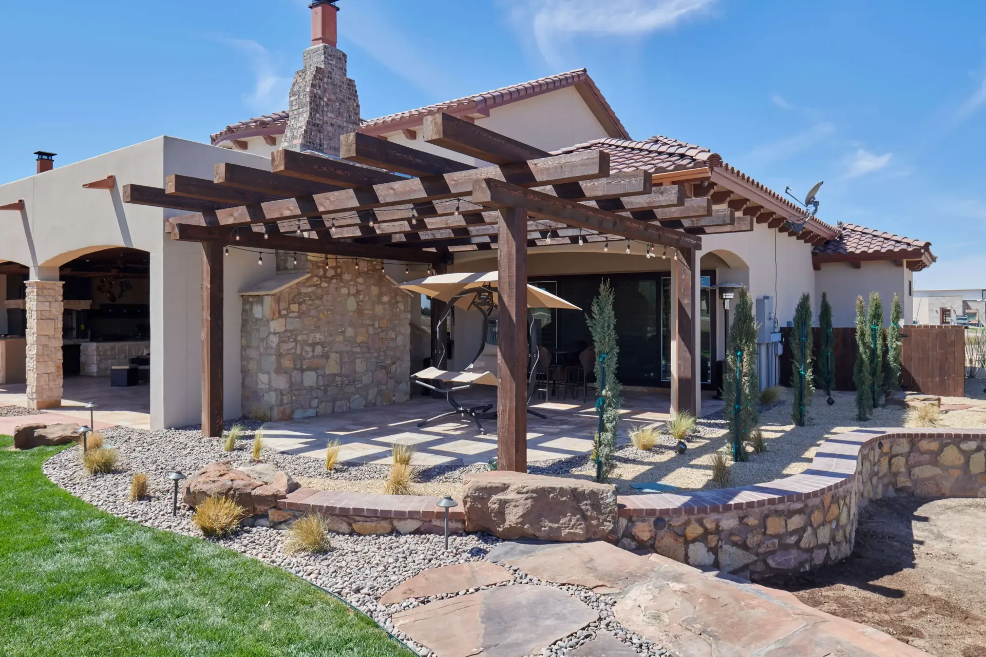 Backyard patio with pergola, stone wall, and landscaping under a sunny blue sky.
