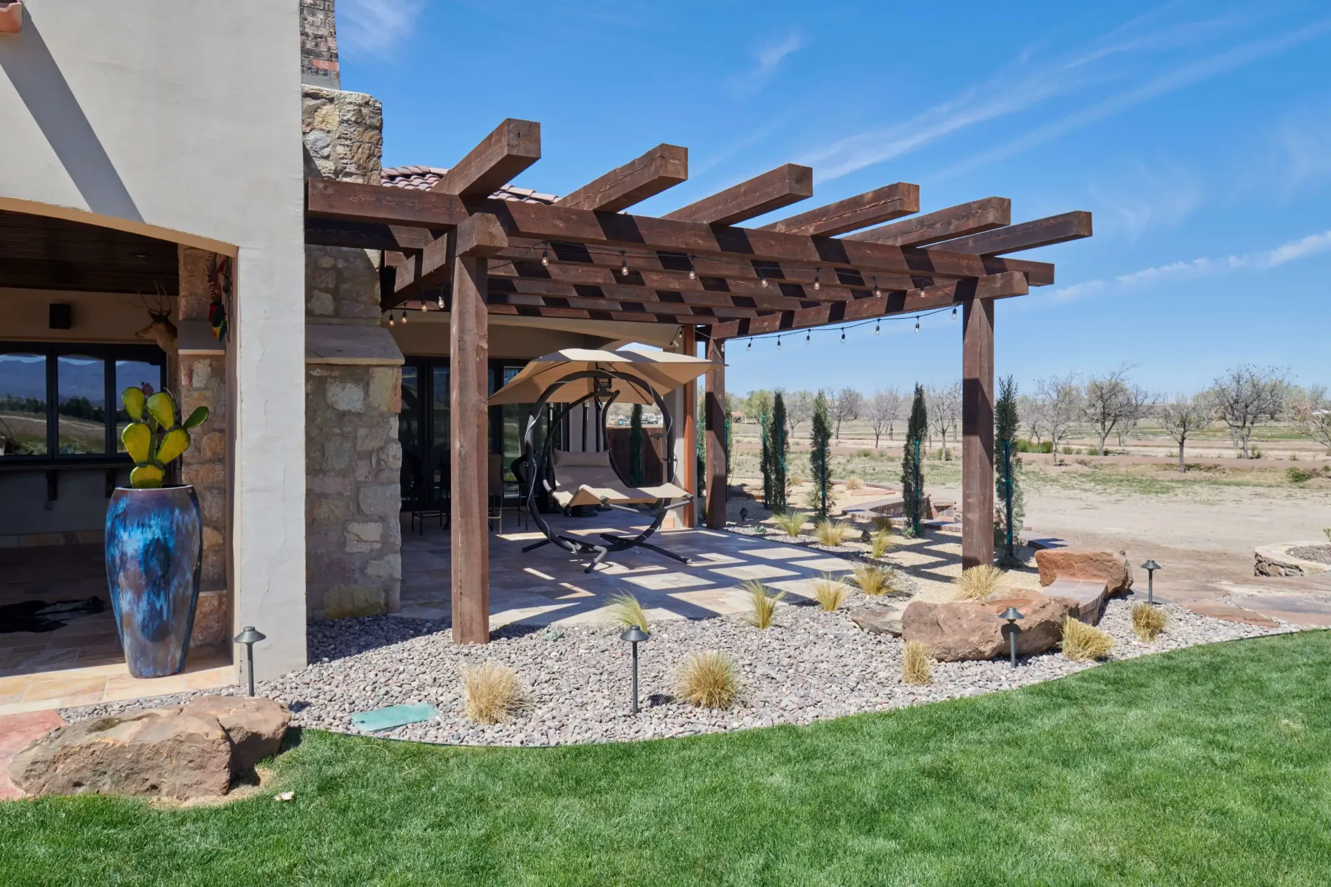 Wooden pergola over a patio. Landscape includes grass, gravel, and small plants. Blue vase next to house.