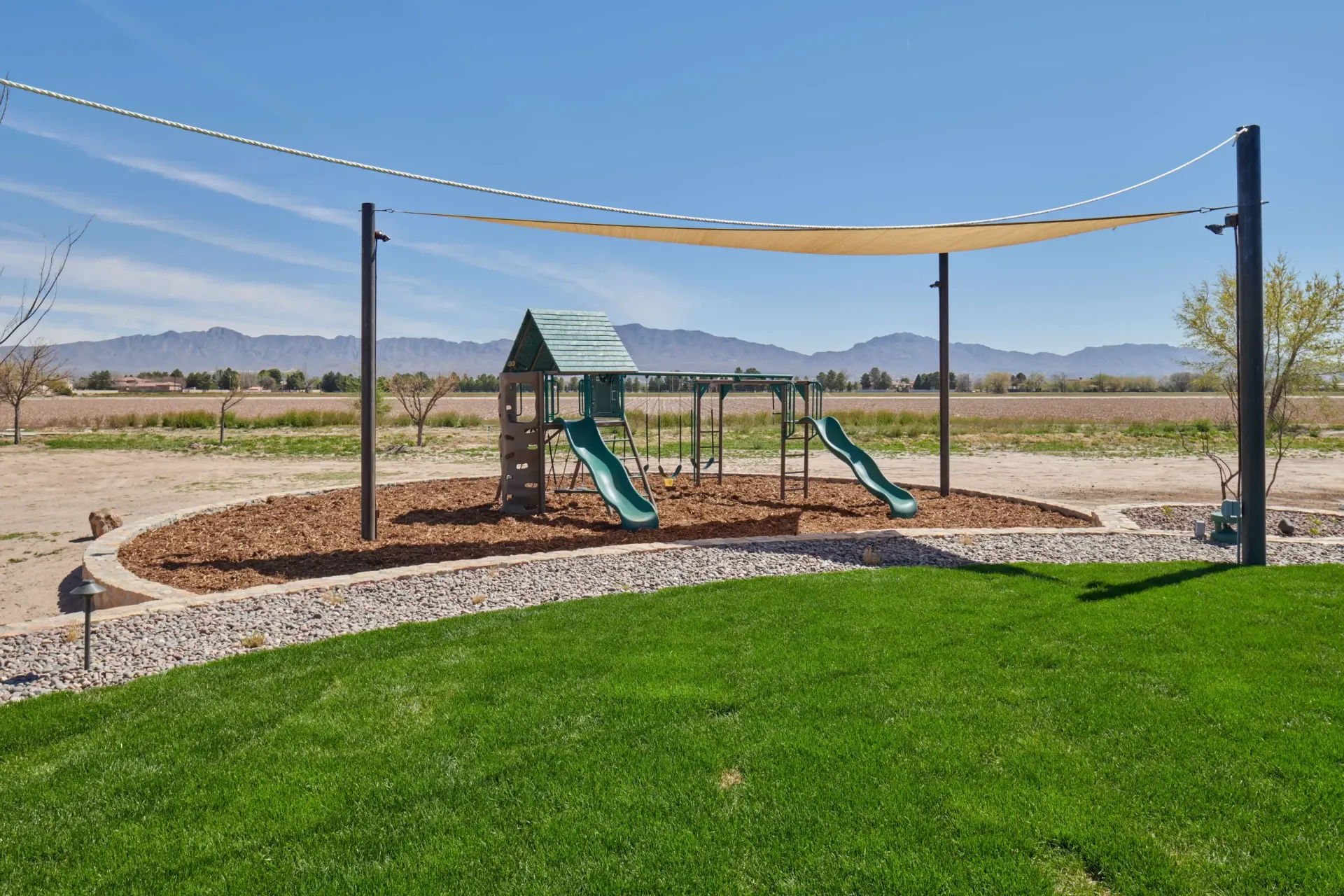 Playground with slides and swings, covered by a sunshade, on a grassy area. Mountains in the background.