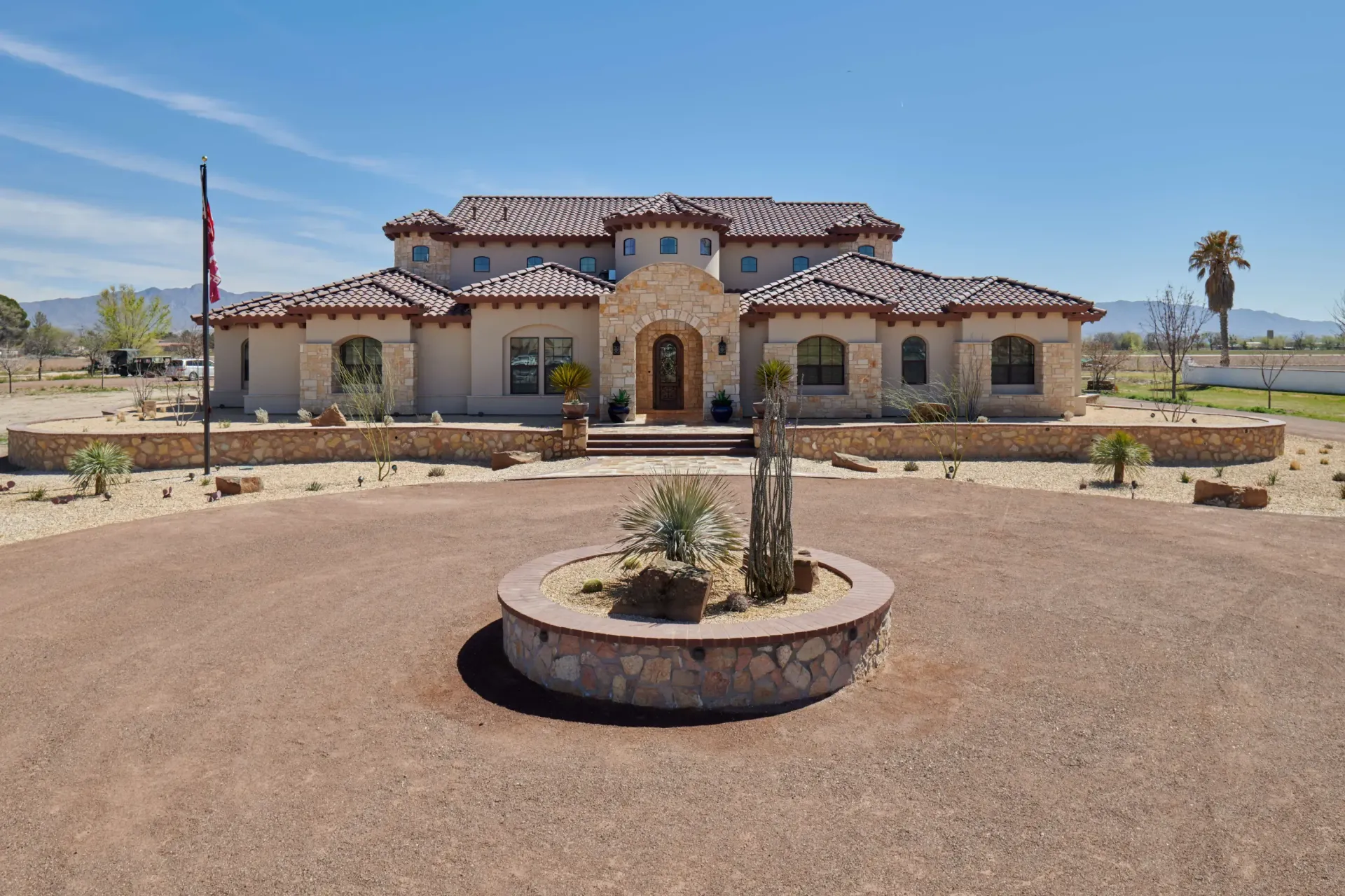 Beige stucco house with tile roof, circular driveway, desert landscaping under a blue sky.