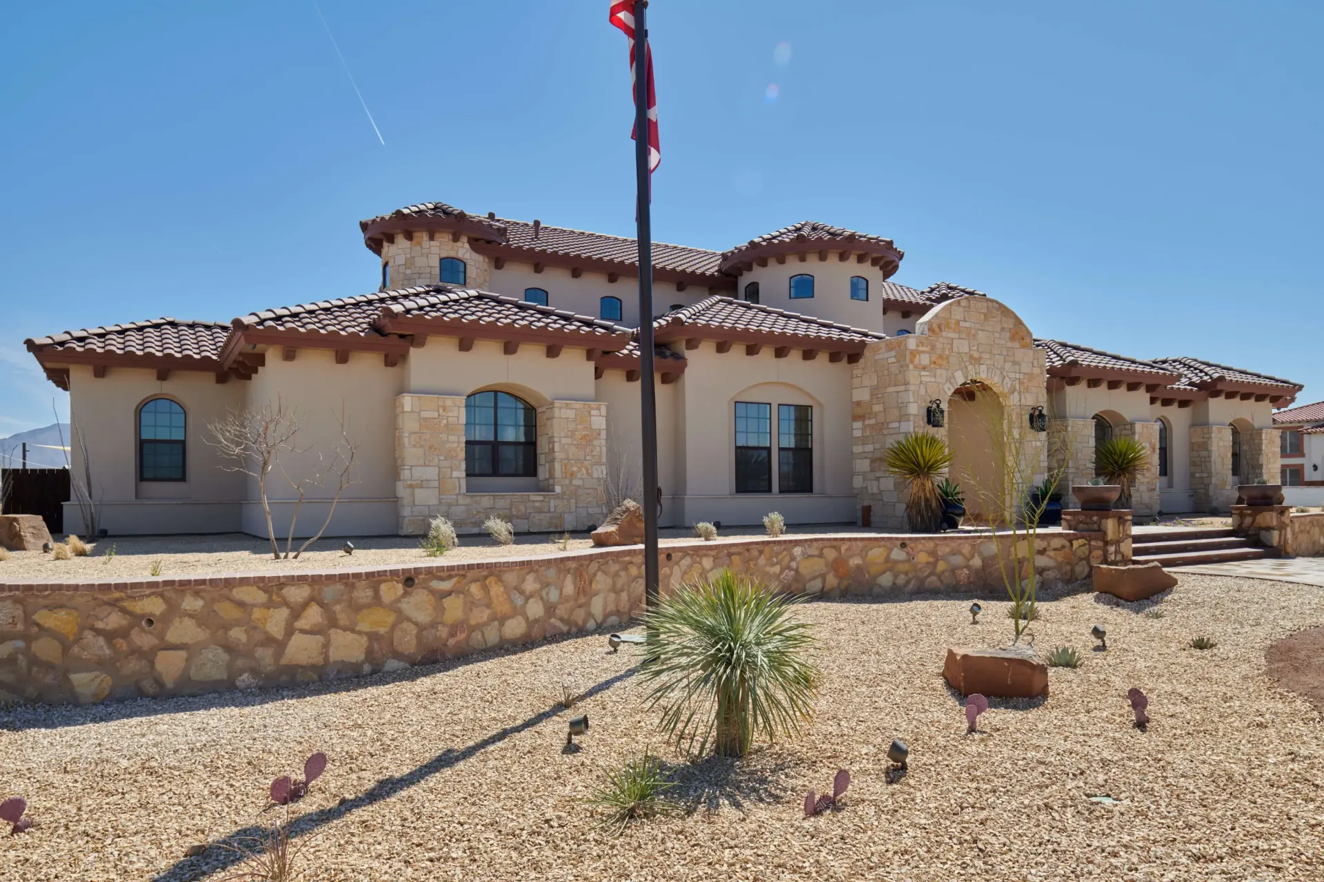 Stucco house with red tile roof, stone walls, and flag pole against a clear blue sky.
