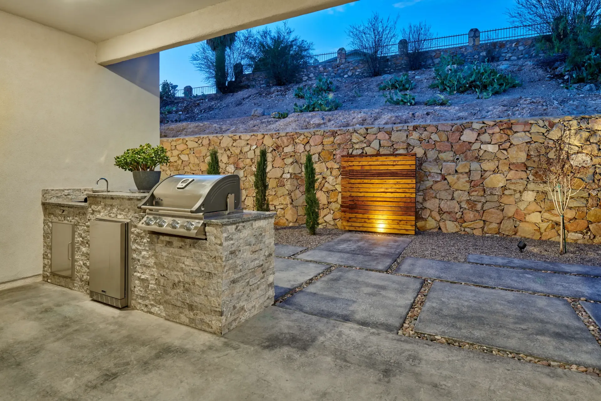 Outdoor kitchen with stone grill, cabinetry, and wooden wall feature; concrete patio with rock accents.