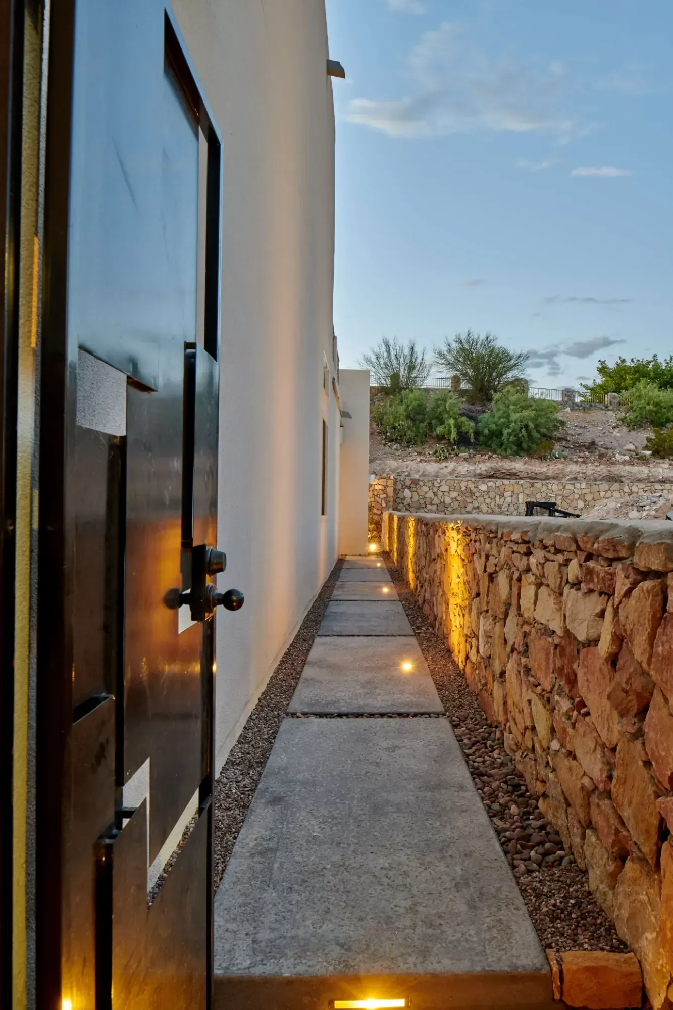 Narrow walkway with embedded lights between a white wall and a stone wall under a dusk sky.