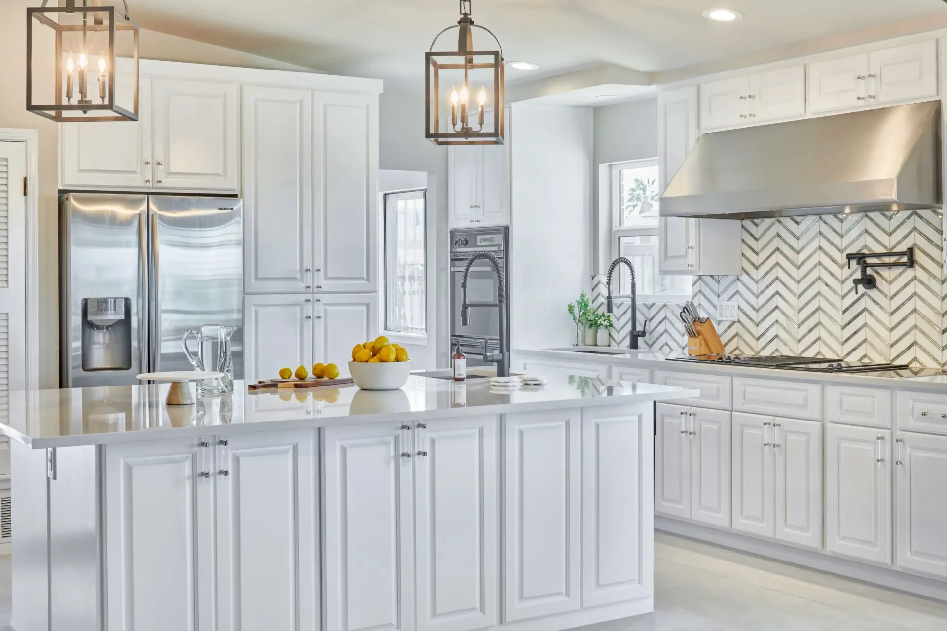 White kitchen with island, cabinets, stainless appliances, herringbone backsplash, and pendant lights.