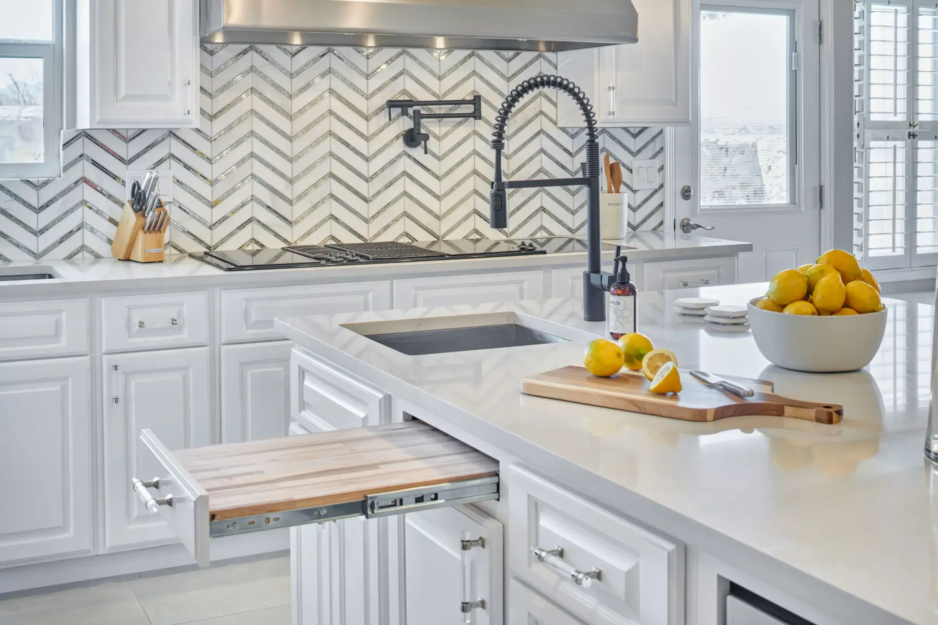 White kitchen with a pull-out cutting board. Island with sink, lemons, and herringbone backsplash.