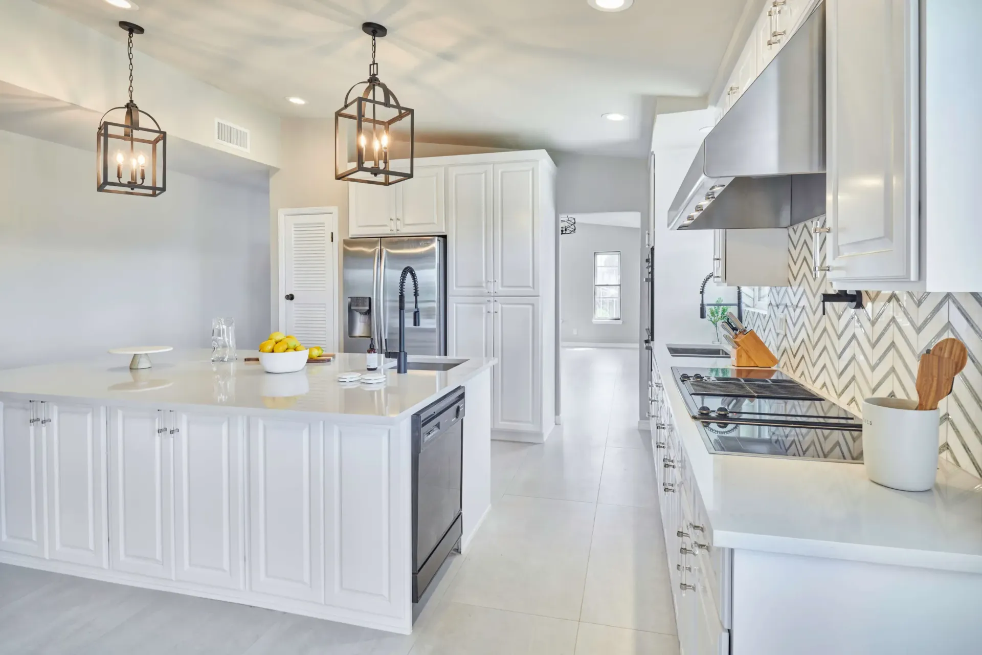 White kitchen with island and pendant lights. Stainless steel appliances and herringbone backsplash.