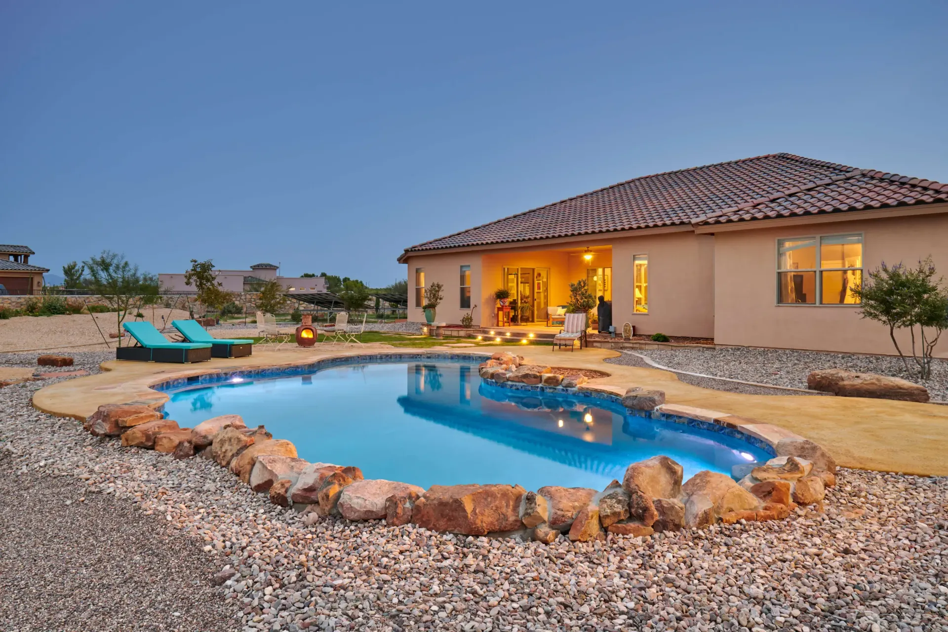 A backyard pool surrounded by rocks, next to a beige house with a terracotta roof.