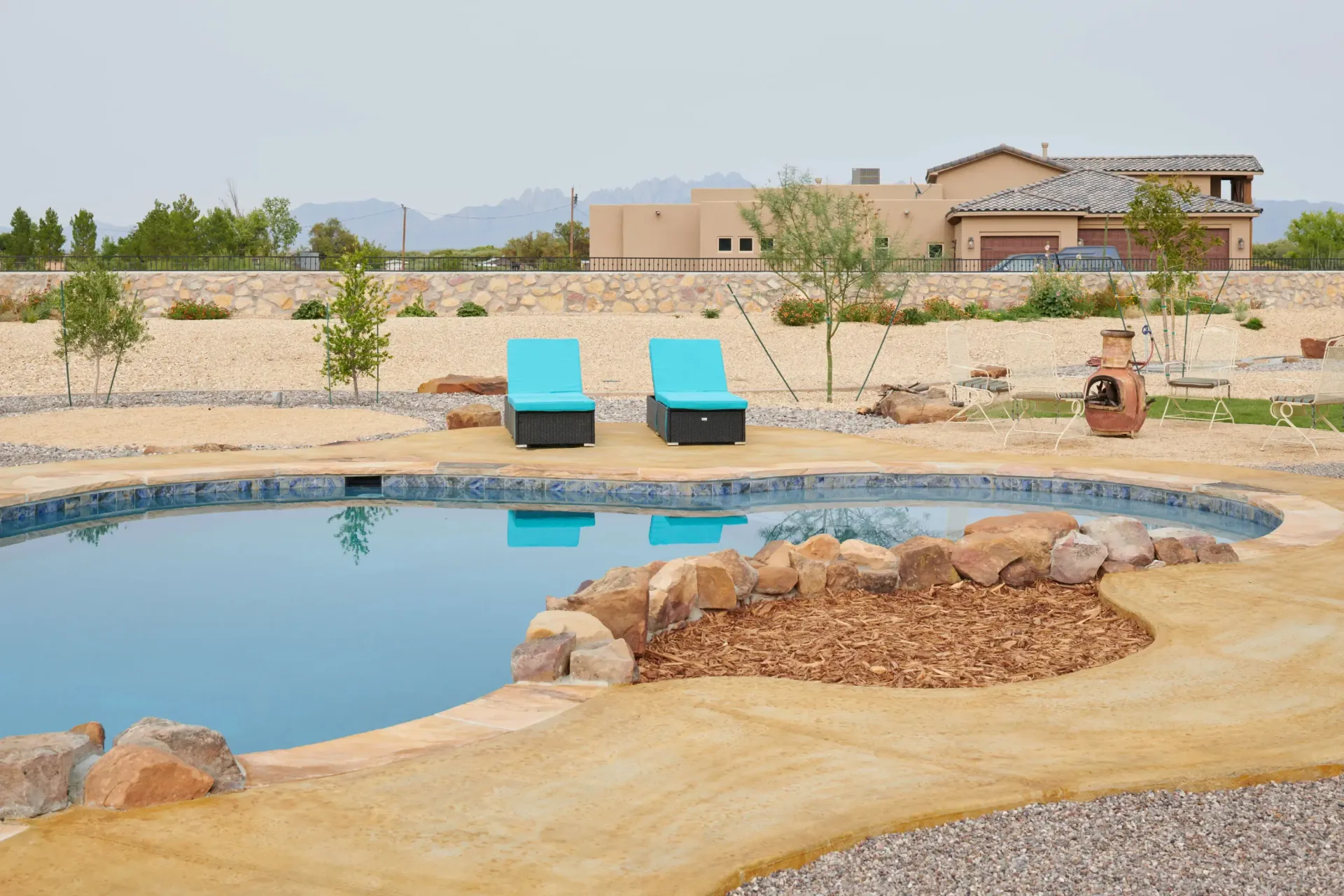 Two blue lounge chairs by a pool in a desert landscape; house in background.