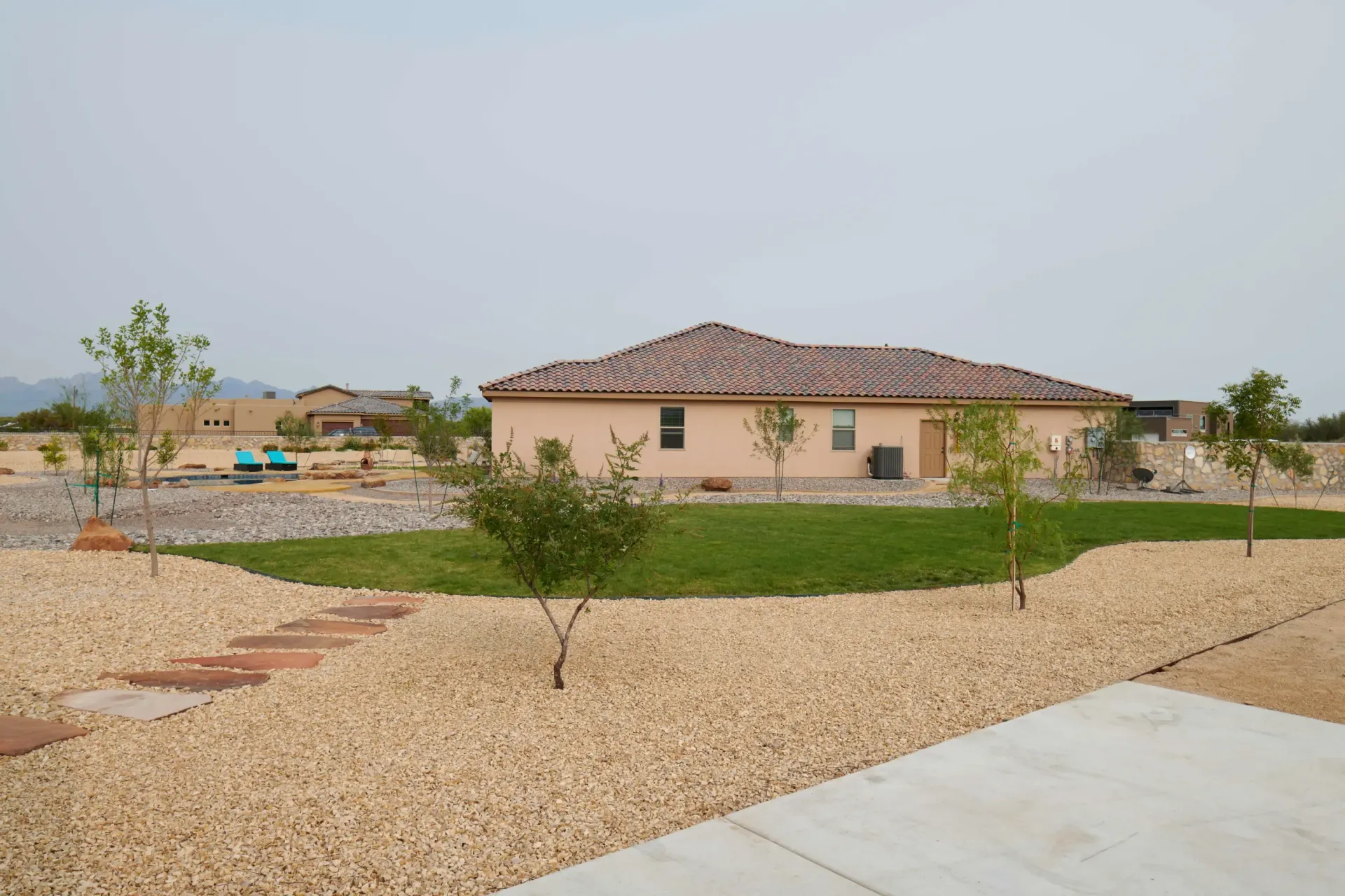 Tan house with a tiled roof, green lawn, and gravel landscaping under a hazy sky.