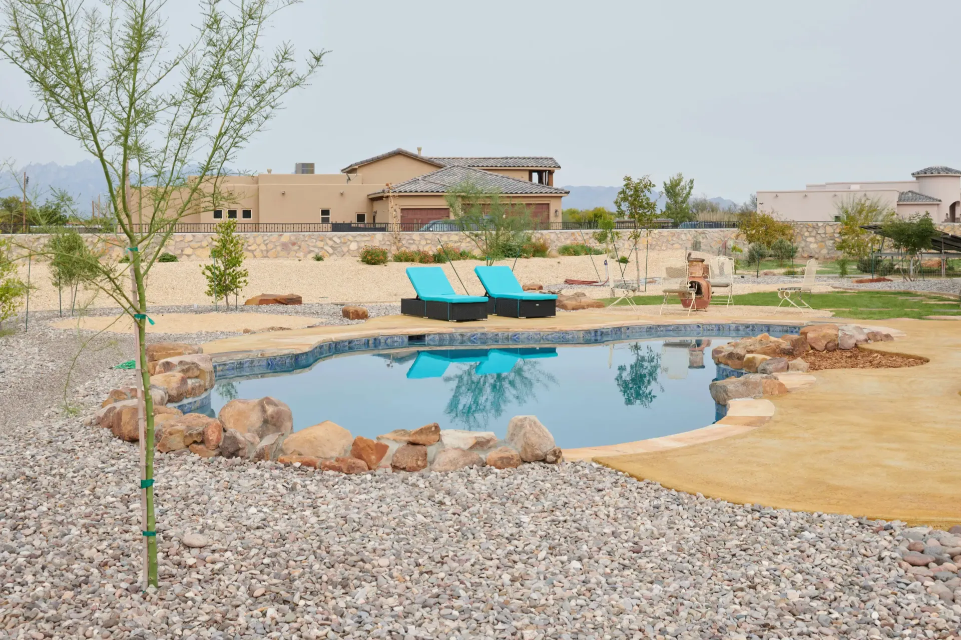 A kidney-shaped swimming pool with blue water and two teal lounge chairs. Rocky landscaping, brown home in the background.