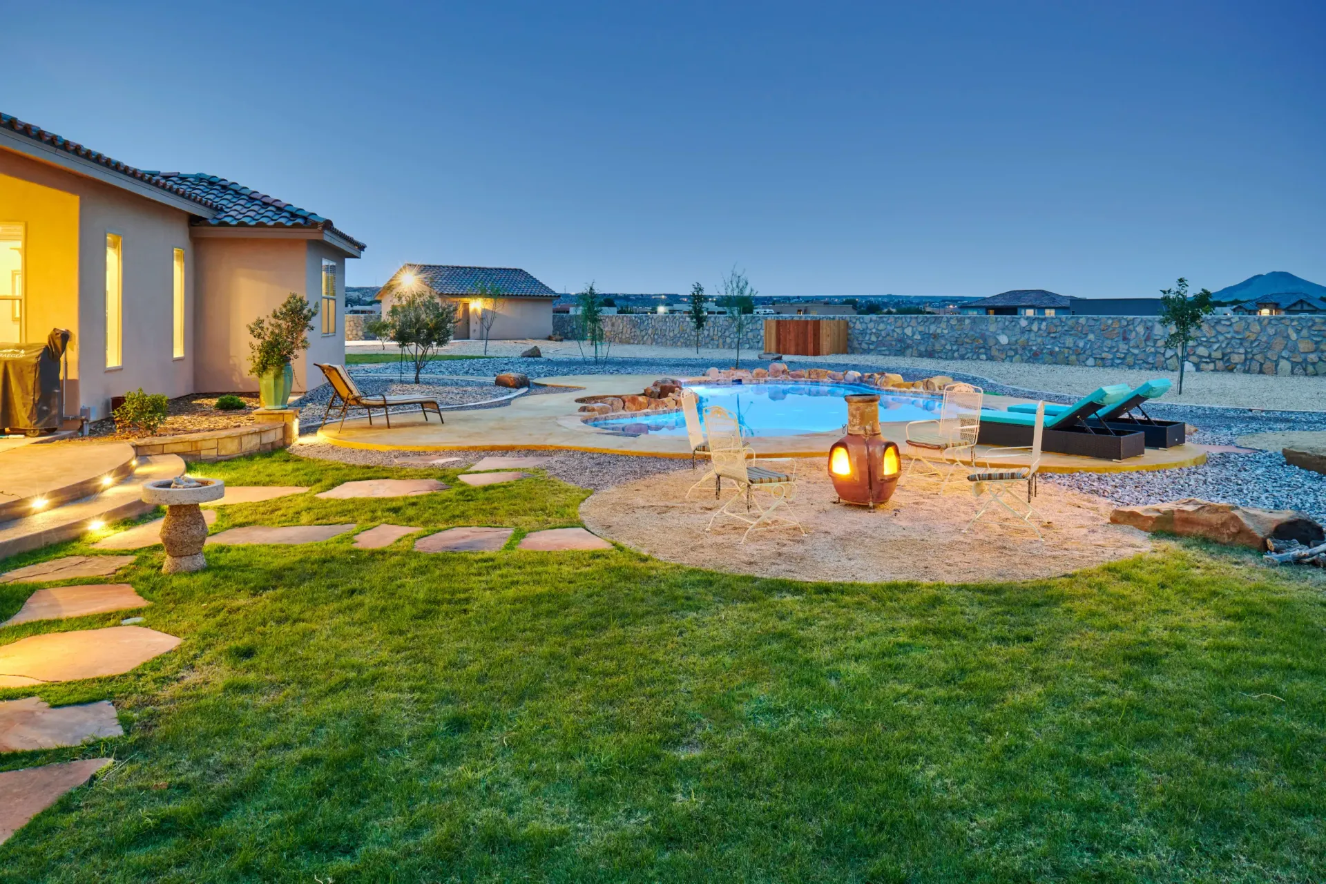 Backyard with a pool, lounge chairs, and landscaping at dusk; house with lights on.