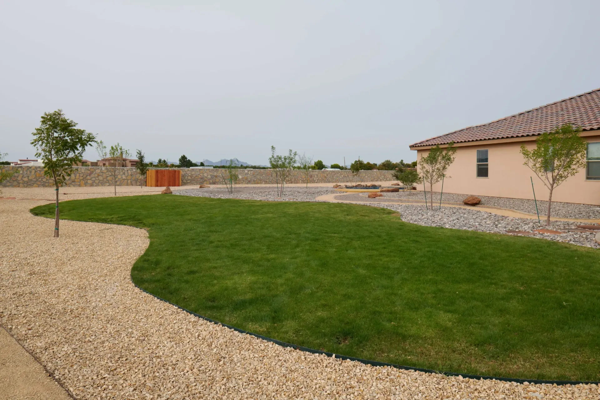 Lush green lawn with curving borders of beige gravel, small trees, and a peach-colored house under a hazy sky.