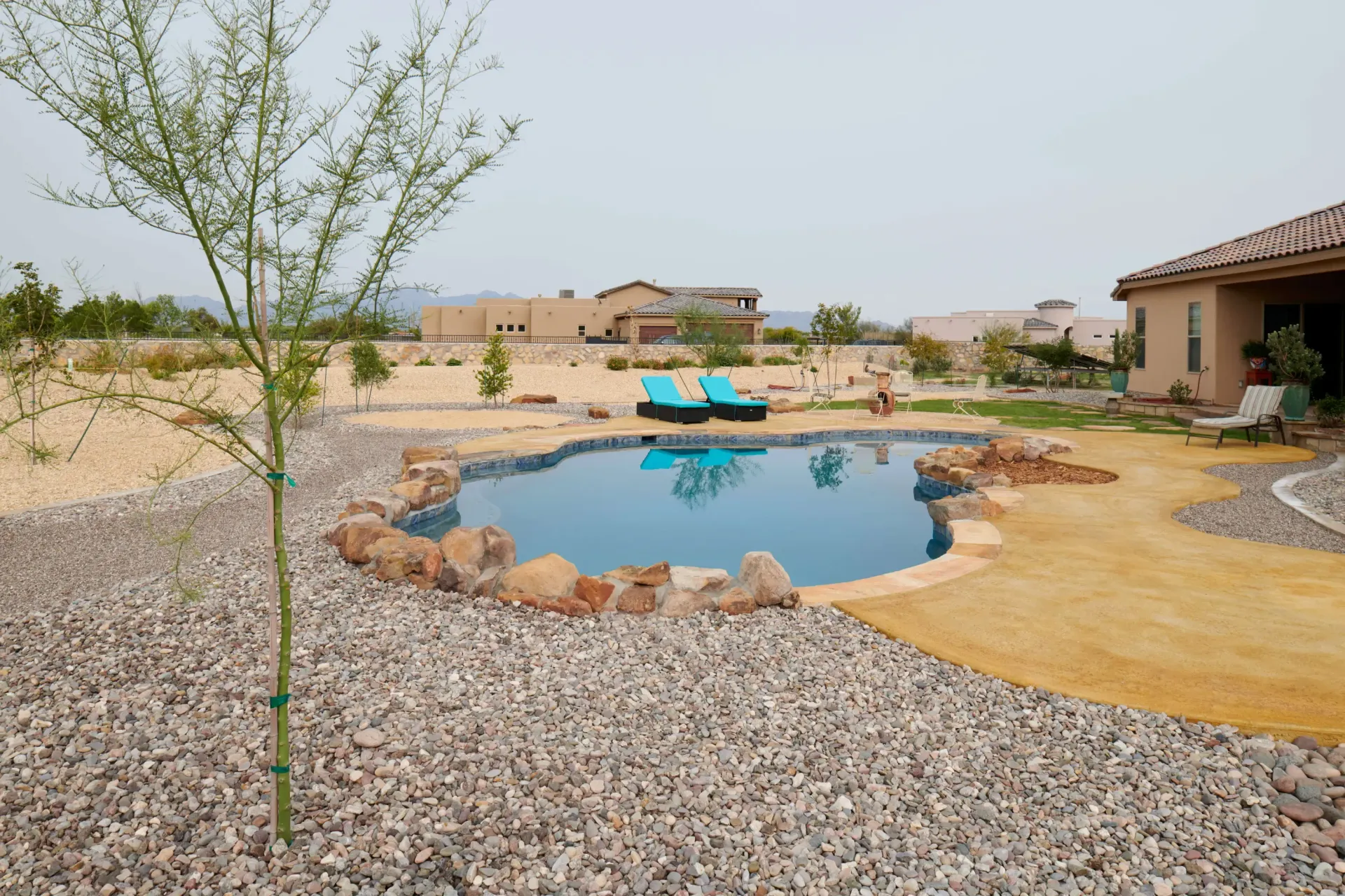 Pool with blue water and stone border, surrounded by gravel and a few trees, with houses in the background.